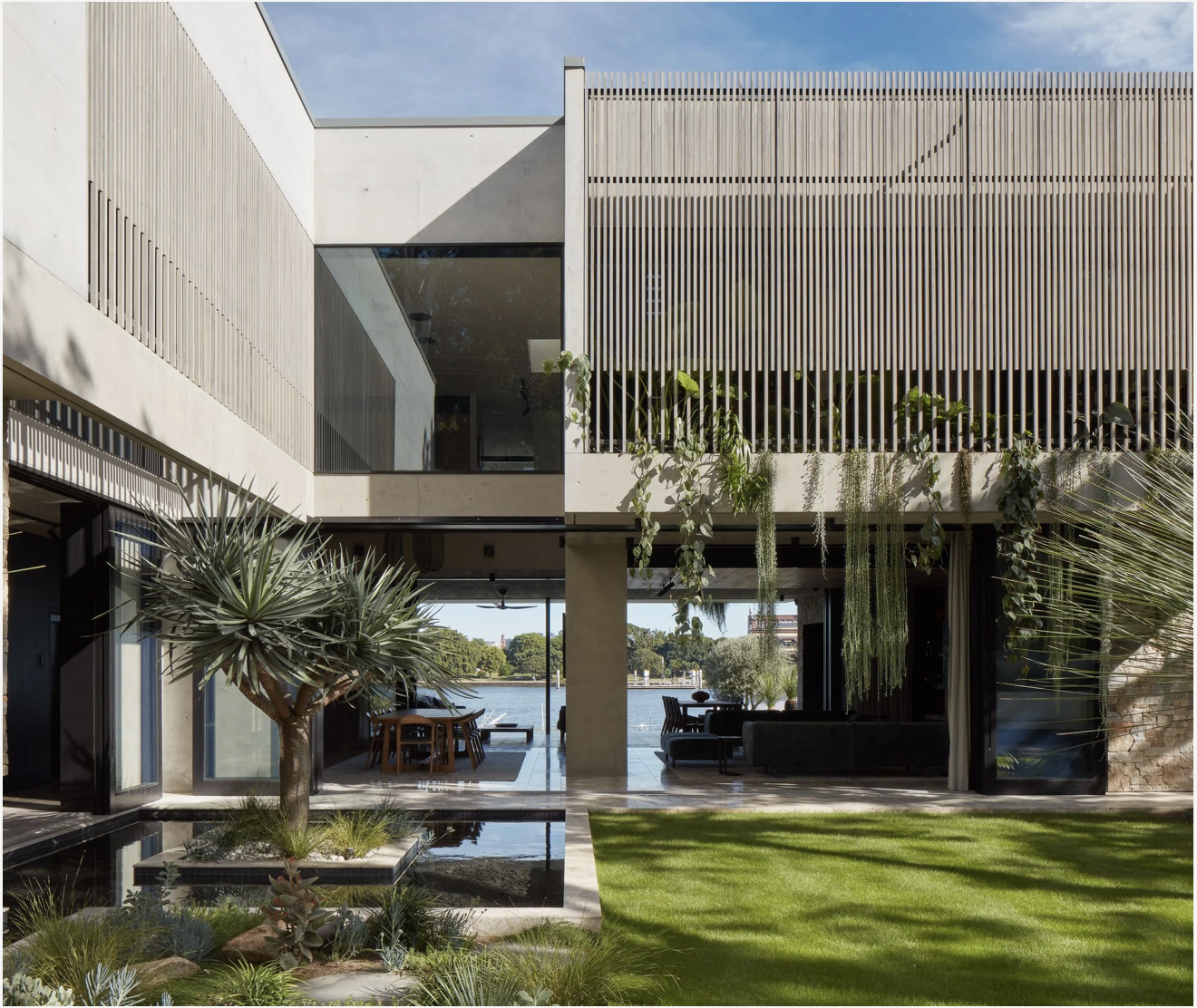 Modern house with large glass windows, outdoor sitting area, water feature, lush green garden, and hanging plants, with a river view in the background.