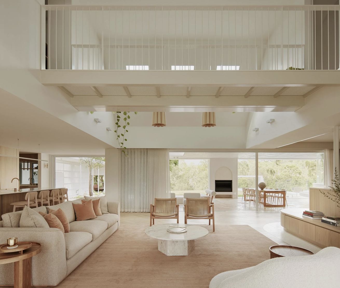 Bright, minimalist living room with beige and wood tones, large sofa with pink pillows, round marble coffee table, armchairs, modern fireplace, and large windows with views of greenery.