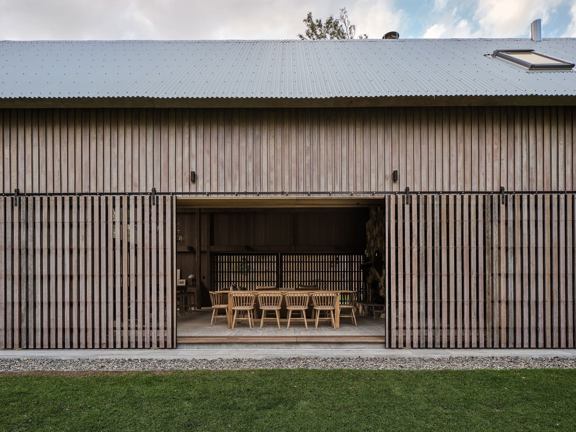 Wooden house with sliding barn doors open to outdoor dining area with wooden table and chairs, green grass in front, gravel edge, cloudy sky above.