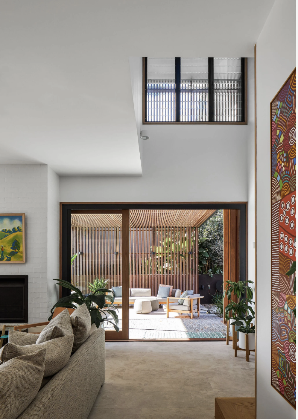 Living room with sliding glass doors opening to an outdoor patio with wooden slats, garden, and seating area, inside on the left are light-colored couches with green plants nearby, art on the walls, and large windows letting in natural light.