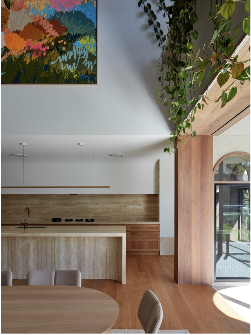 Interior view of a modern kitchen and dining area with wooden accents, a hanging abstract colorful painting, and a large window with a view of greenery outside. There is also a leafy plant hanging from the ceiling.