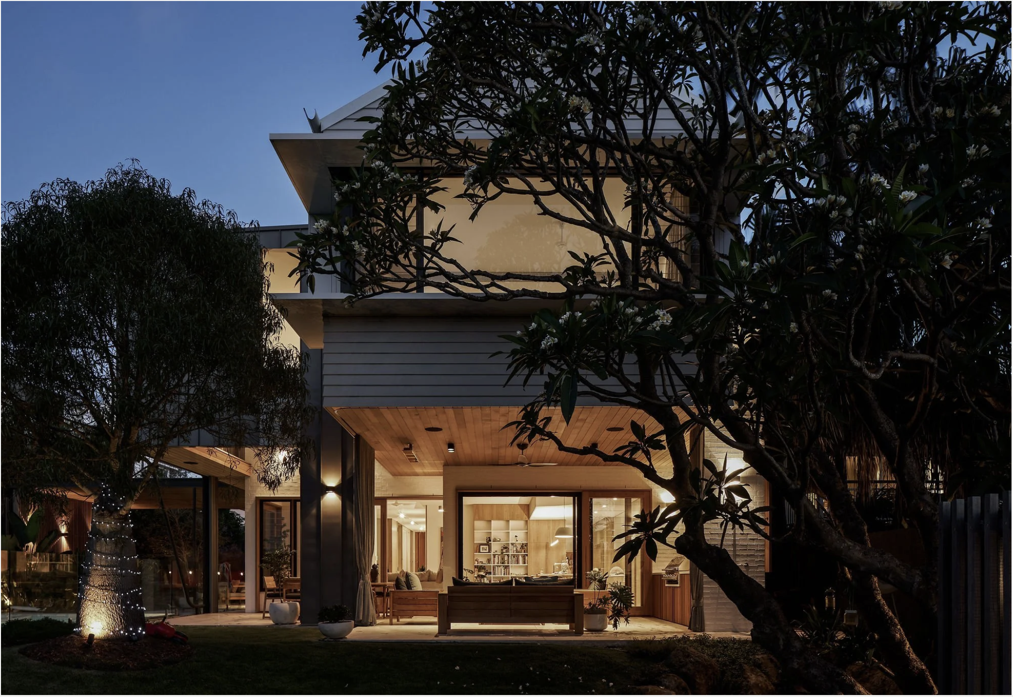 Modern two-story house illuminated at dusk, with large glass windows showcasing a cozy interior, surrounded by trees and outdoor furniture.