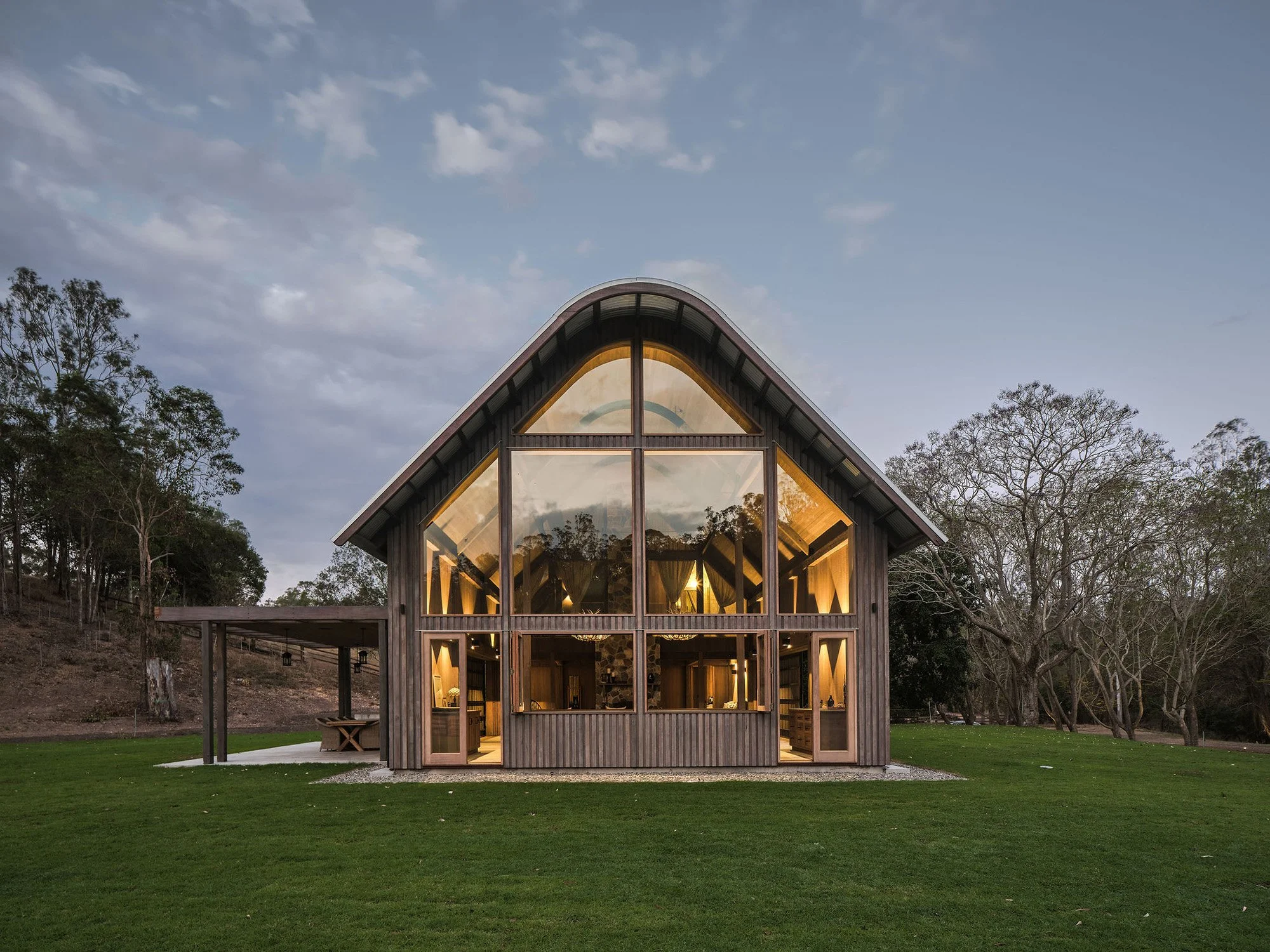 A modern house with wooden exterior and large glass windows, situated on a grassy lawn with trees in the background, during dusk.
