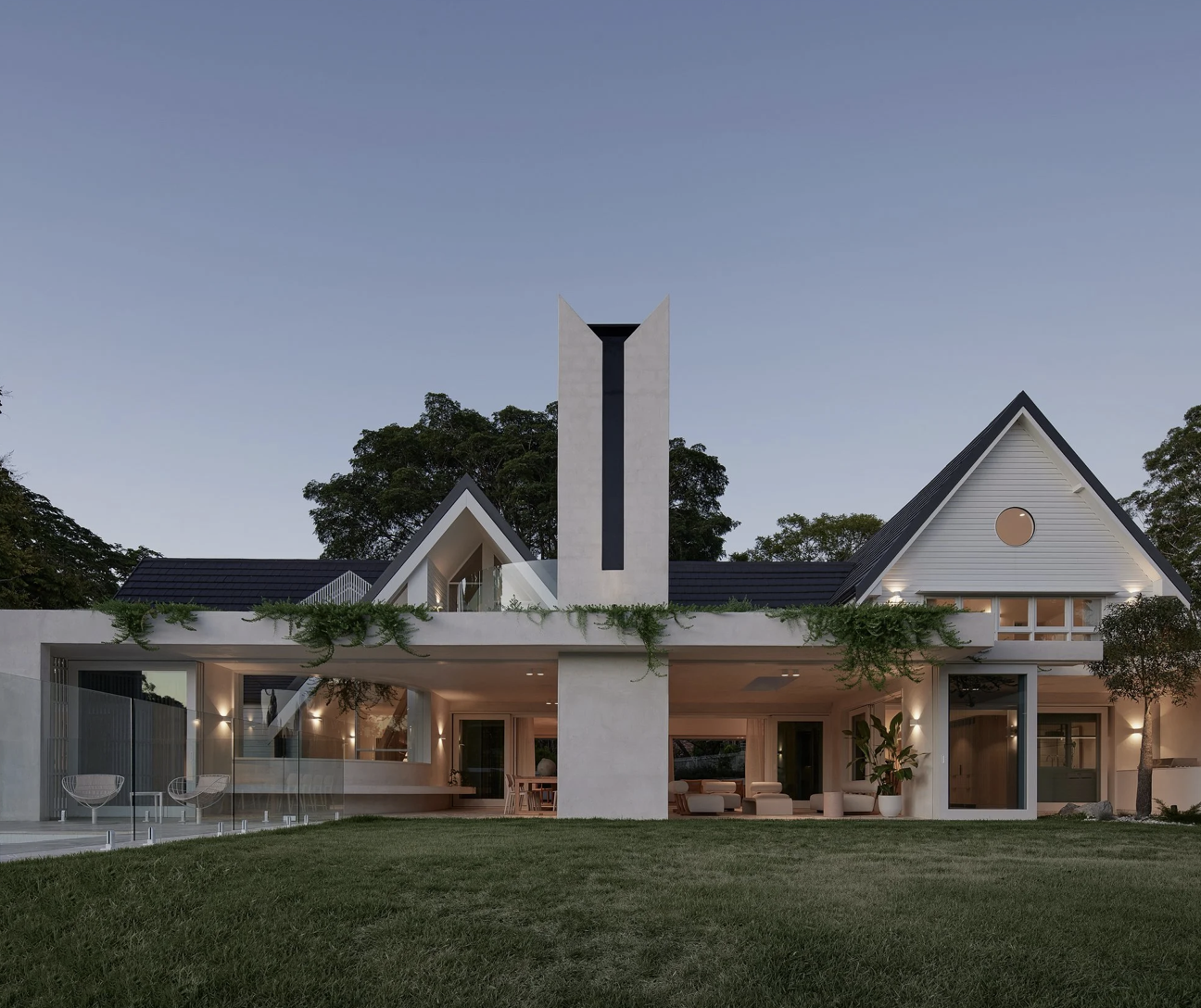 Modern house with white exterior, black roof, and large windows, surrounded by greenery and trees, at dusk.