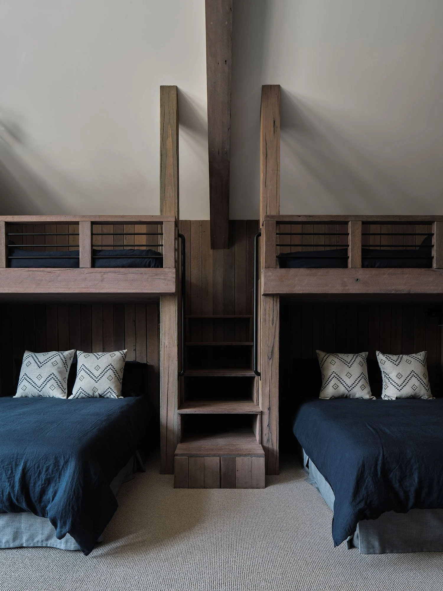 A bedroom with two single beds and two upper bunk beds, all made of dark wood, with decorative pillows on the main beds, in a room with sloped ceiling.