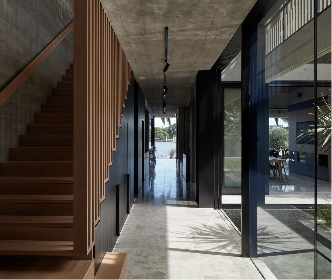 Interior view of a modern house with open hallway, wooden staircase on the left, glass sliding doors on the right, and outdoor patio with water view in the background.