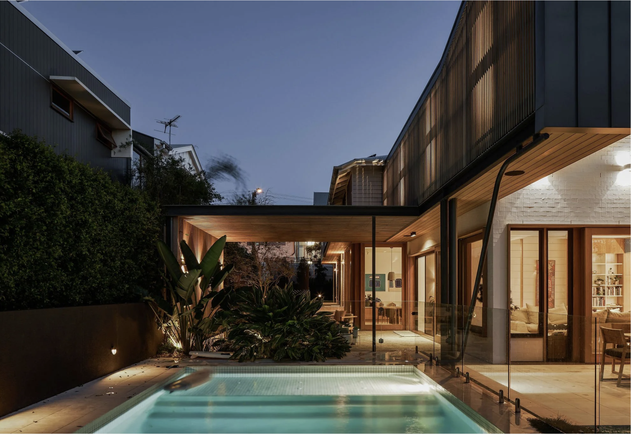Modern house with glass doors and wooden accents, backyard with swimming pool and garden at dusk.