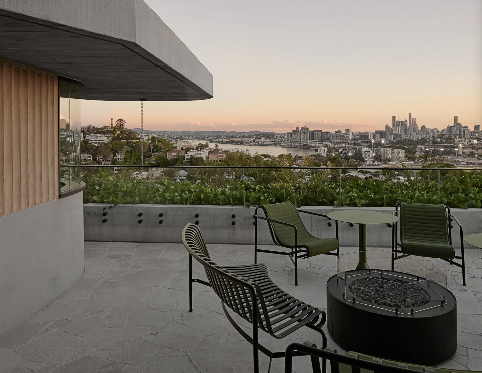 An outdoor patio with modern chairs around a fire pit, overlooking a cityscape with high-rise buildings and a river at sunset.