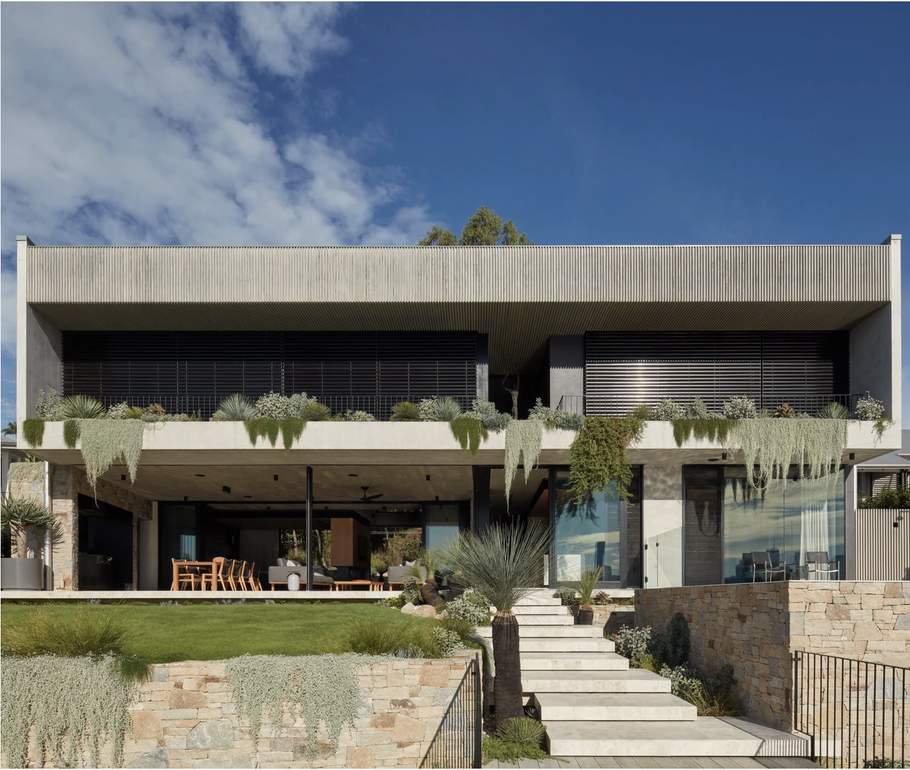 Modern multi-story house with a flat roof, large glass windows, and a landscaped front yard with plants, grass, and stone steps leading up to the entrance.