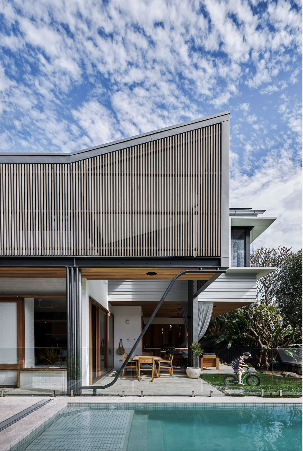 Modern house with wooden and metal exterior, swimming pool in foreground, child riding bike on grass, blue sky with scattered clouds.