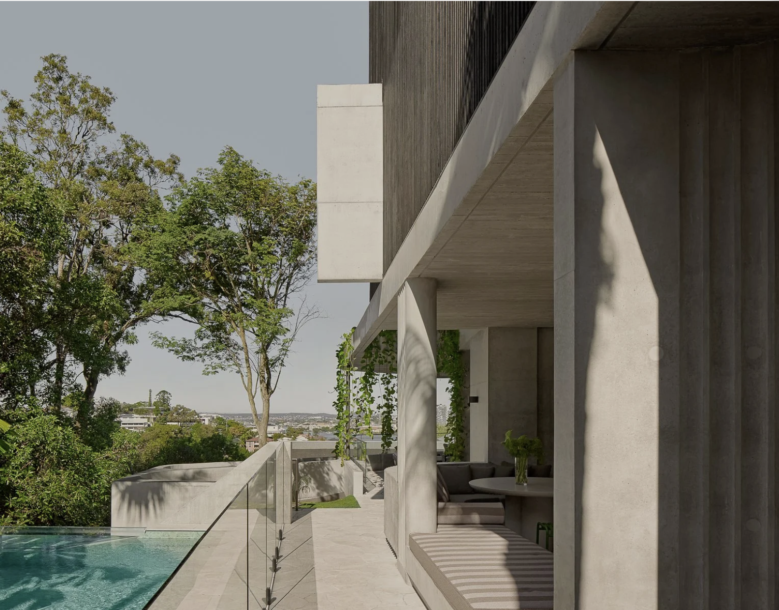 Modern patio with concrete structure, glass railing, outdoor seating with striped cushions, a small table with a vase of flowers, overlooking a swimming pool and trees, under a clear sky.