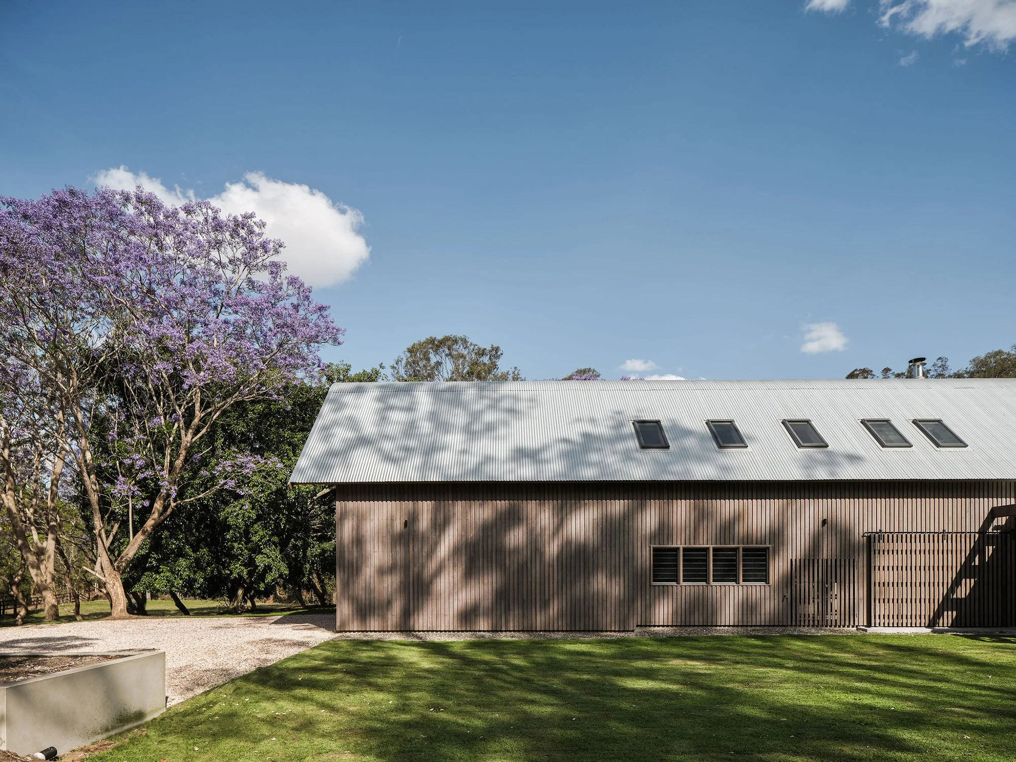 A modern wooden building with a metal roof and multiple skylights. There is a green lawn and a large tree with purple blossoms to the left, against a blue sky with a few clouds.