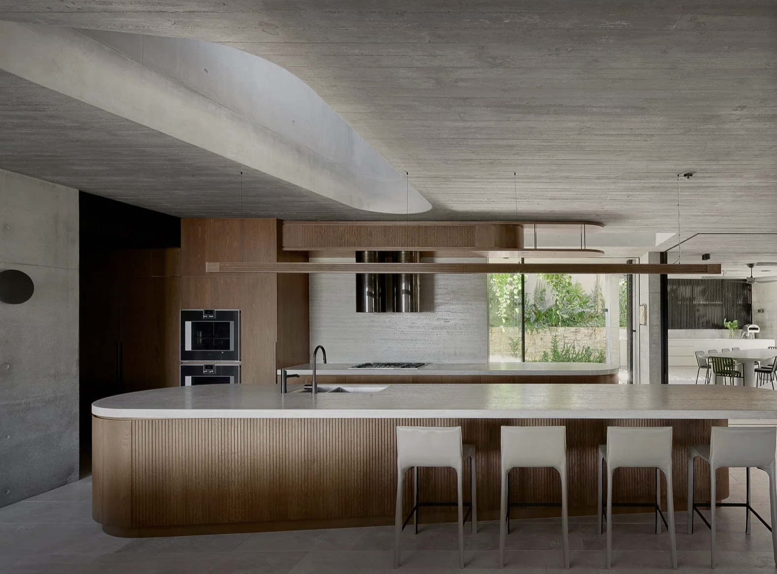 Modern kitchen with curved wooden island, white chairs, black appliances, large window with greenery outside, and minimalist decor.