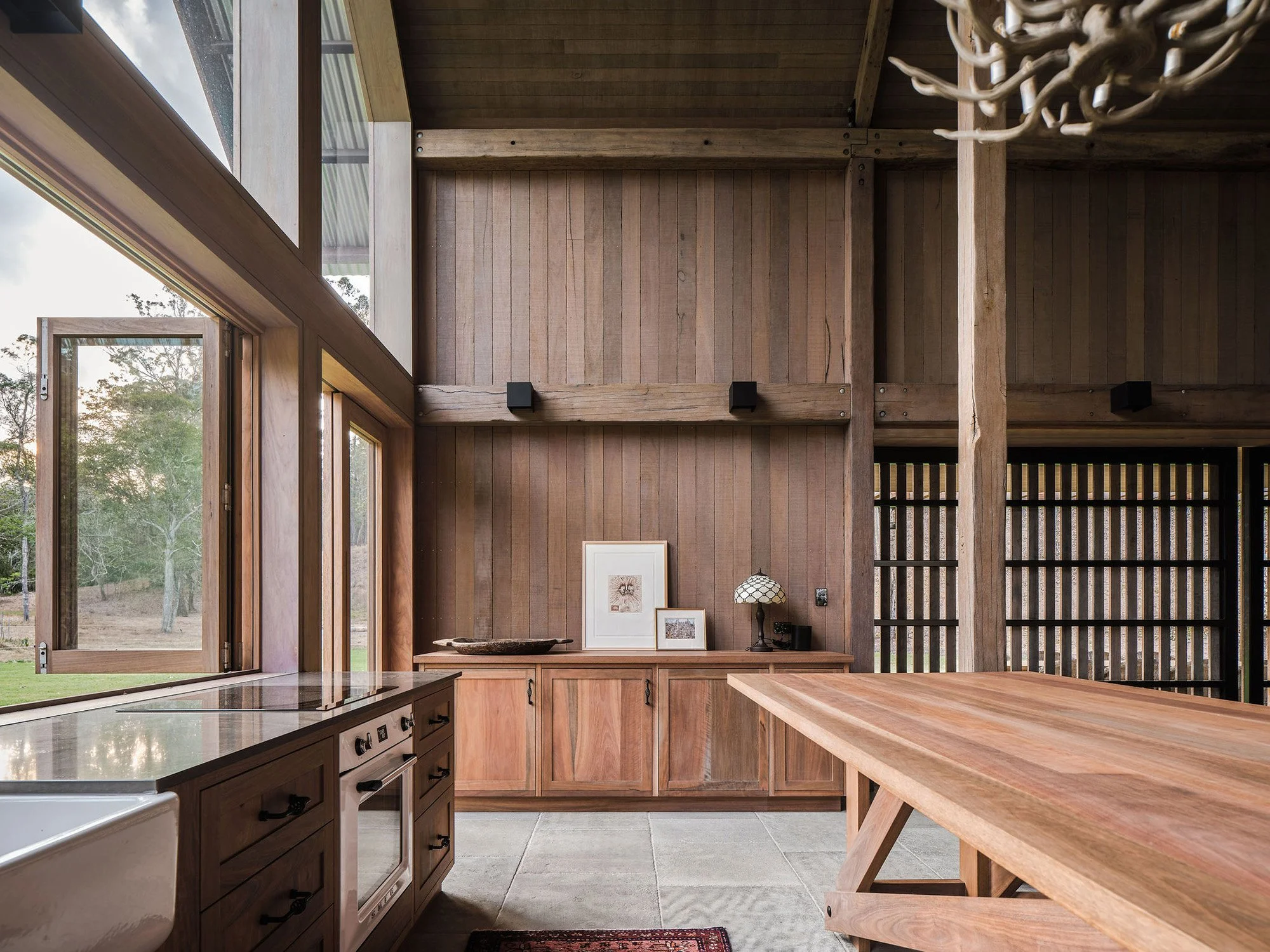 Interior view of a kitchen with wooden walls and cabinetry, large windows showing an outdoor landscape, a wooden dining table, and decorative framed pictures on the sideboard.