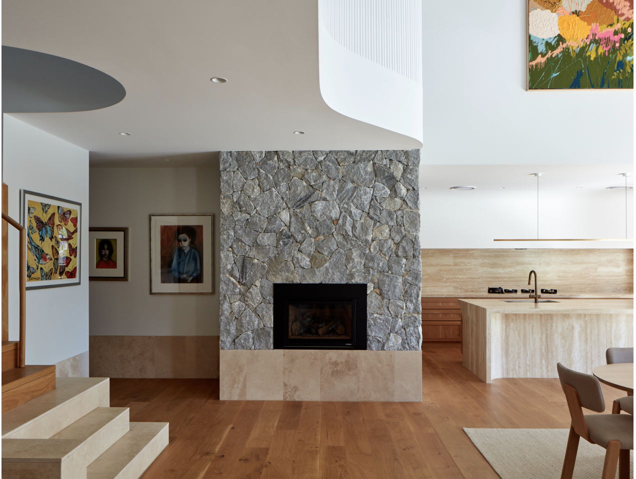 Interior of a modern home featuring a stone fireplace with a glass-enclosed fire, wooden floors, an open kitchen with a beige island, and colorful wall art.