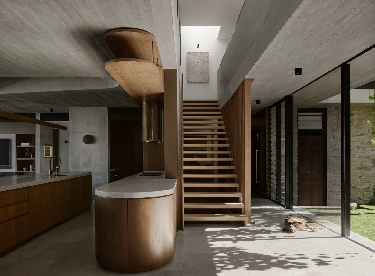 Modern interior of a house with wooden staircase, kitchen with wooden cabinets and marble countertop, and large glass sliding door leading to outdoor grass area with a dog lying in the sun.