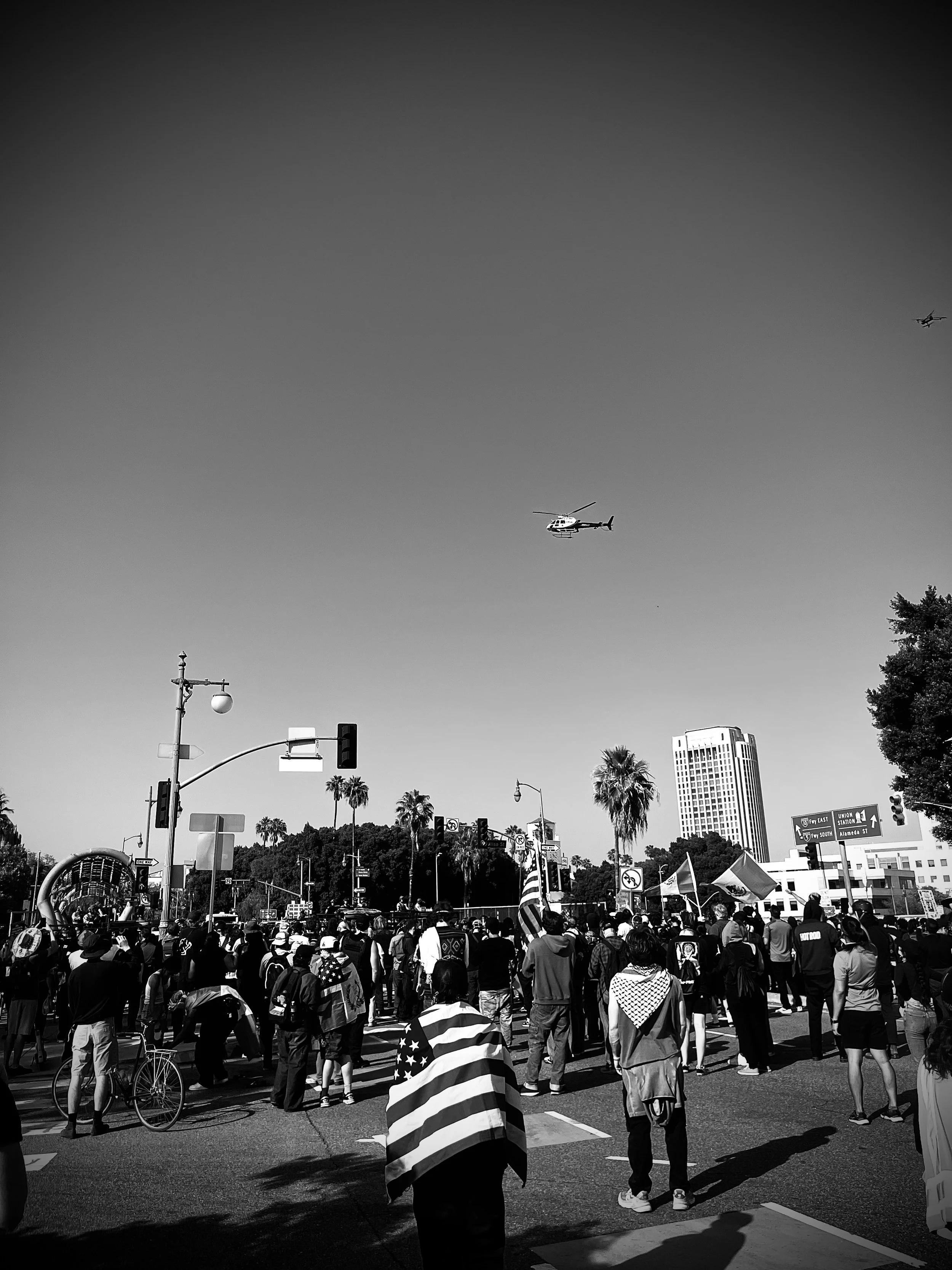 A group of protesters watches as an LAPD helicopter hovers overhead and a line of anti-riot police vehicles stands nearby. (Los Angeles, June 2025)
