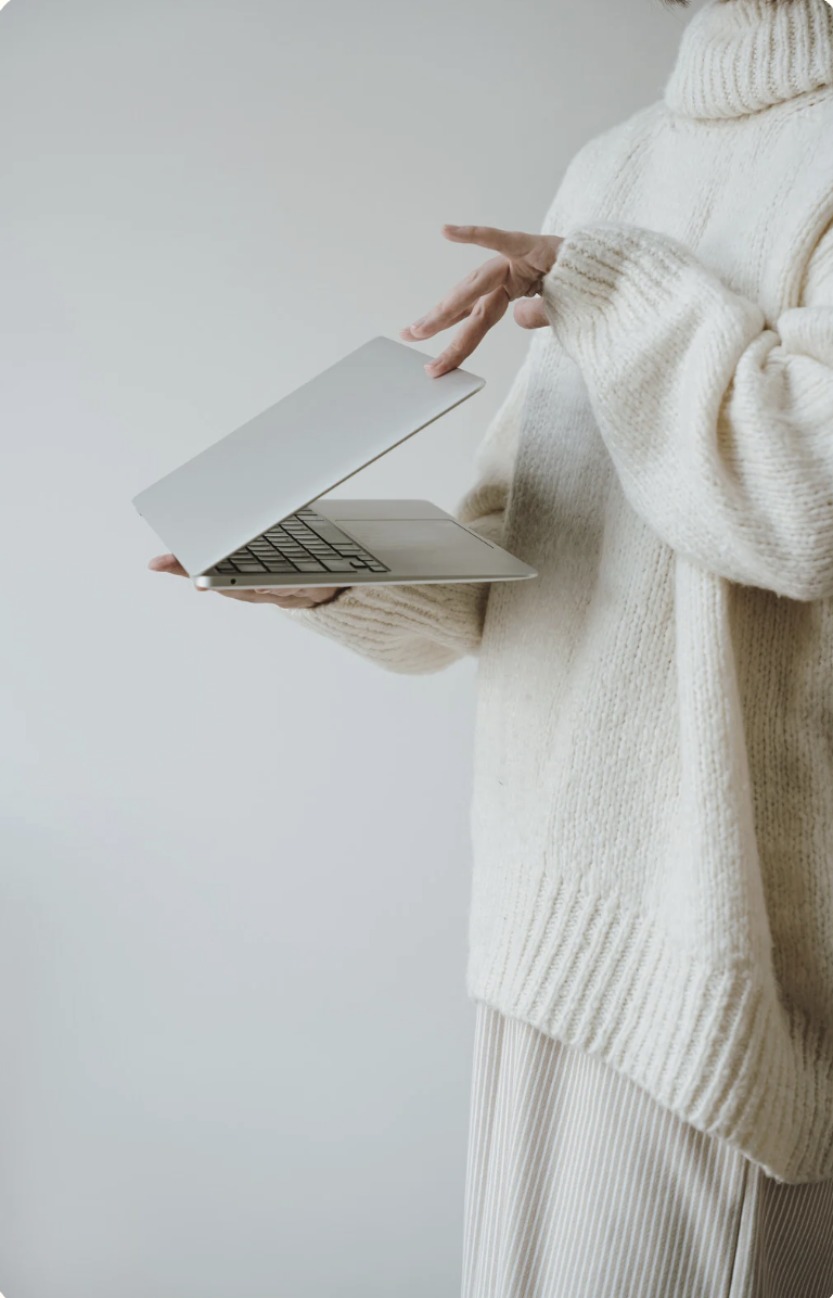 Person in a cream-colored sweater holding a silver laptop with one hand and using the other hand to scroll.
