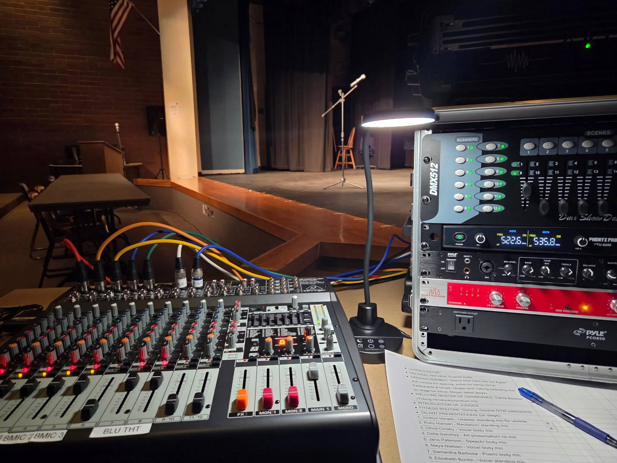 A stage setup with a microphone on a stand, chairs, and a curtain in the background. In the foreground, there is audio equipment including a sound mixer and a rack-mounted audio processor. The scene appears to be prepared for a live performance or event.