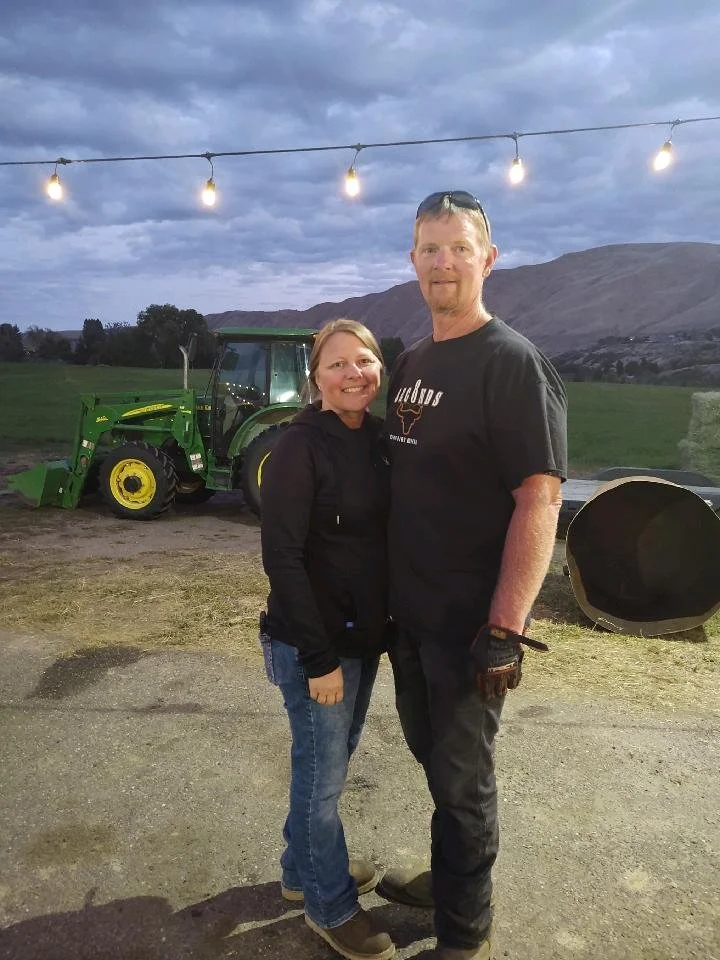 A woman and a man standing outdoors at dusk with a tractor and mountains in the background. They are smiling and facing the camera.