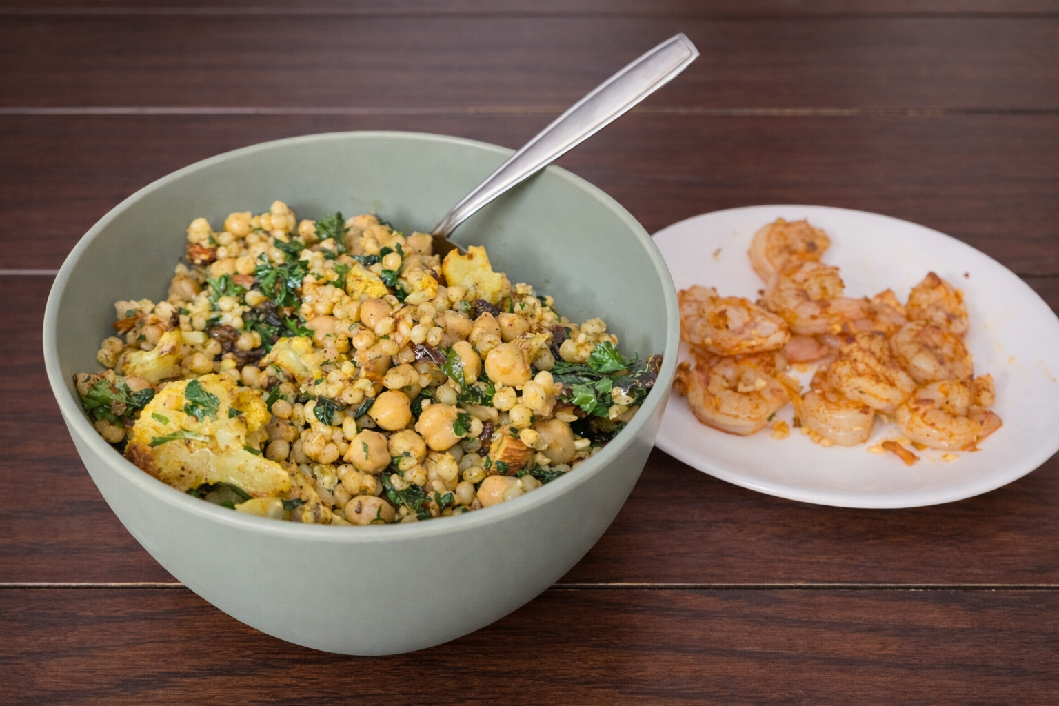 Bowl of cauliflower couscous salad with roasted cauliflower, couscous, chickpeas, and herbs, served alongside lemon-garlic shrimp on a white plate.