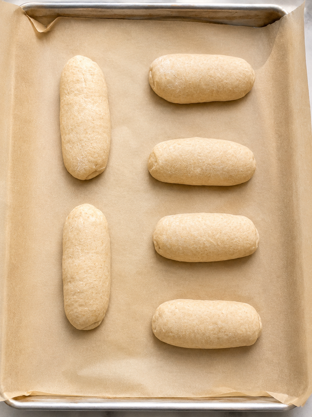 Six shaped whole wheat sub rolls on a parchment-lined baking sheet before baking.