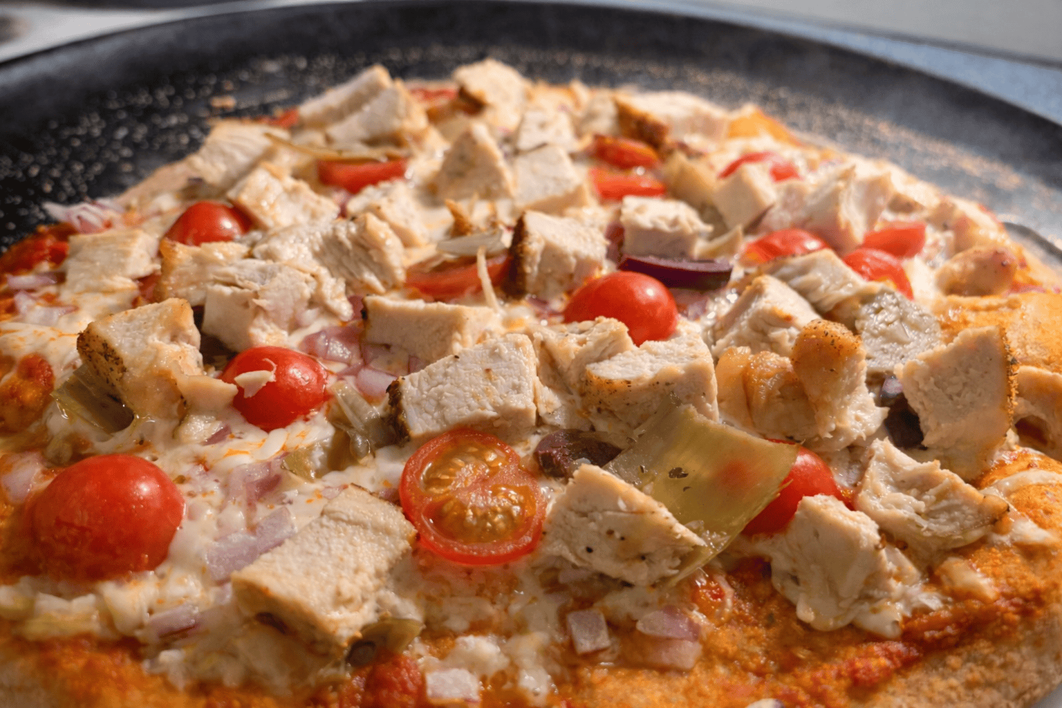Close-up of homemade pizza topped with grilled chicken, cherry tomatoes, red onion, and melted cheese on a crispy crust.