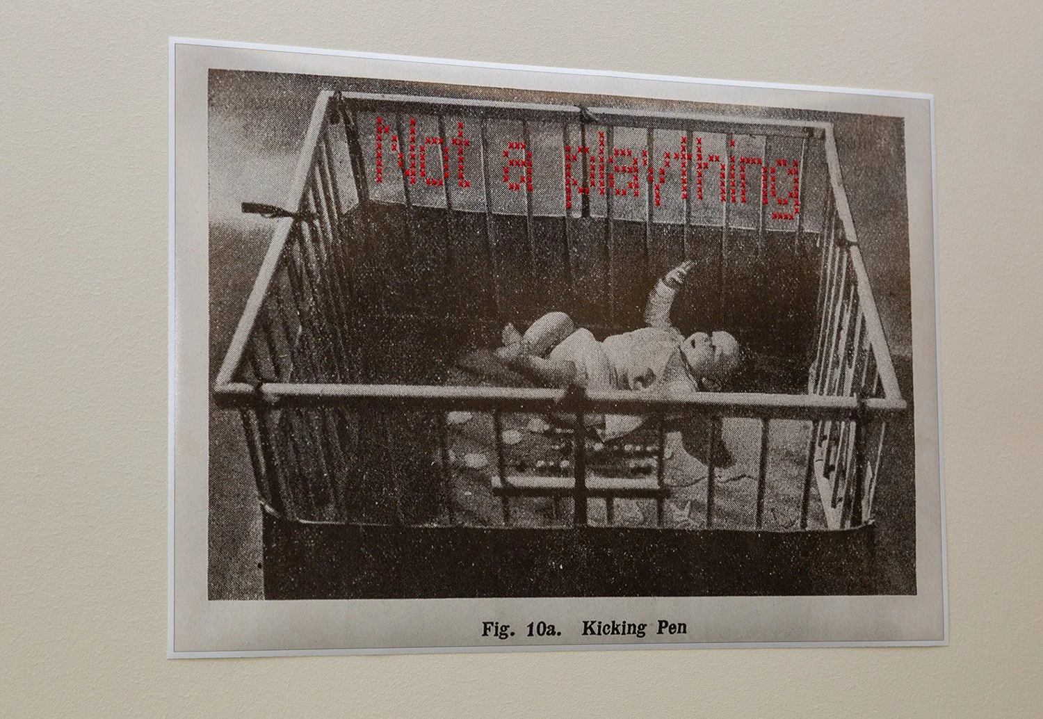 A picture of a baby in a kicking pen from Sir Frederic Truby King's 1913 book 'Feeding and Care of Baby' and the words 'Not a plaything' cross-stitched to play on his controversial ideologies. Image in 1942 revised edition.