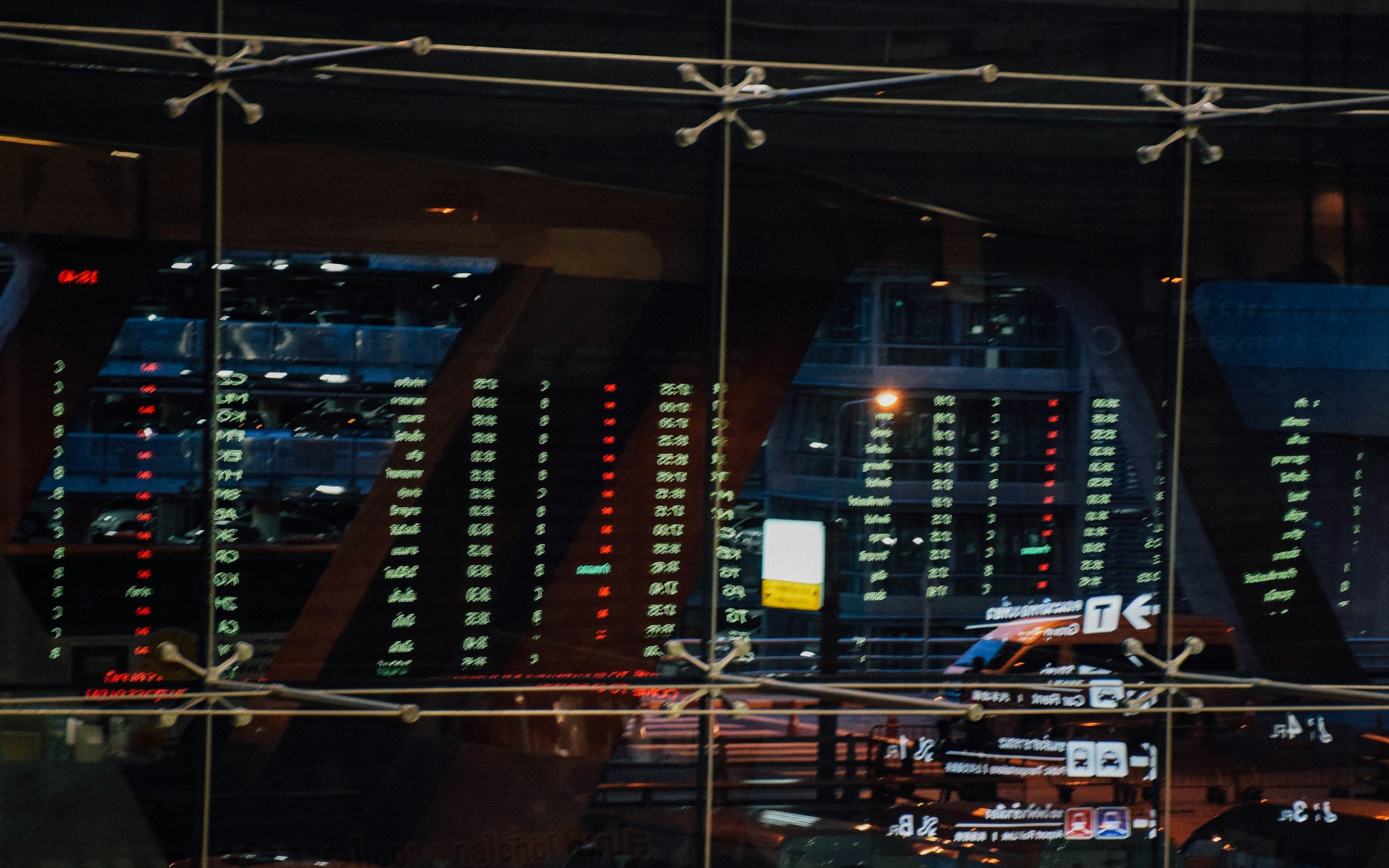 Nighttime airport departure board with illuminated flight details and signs reflected in a glass window.