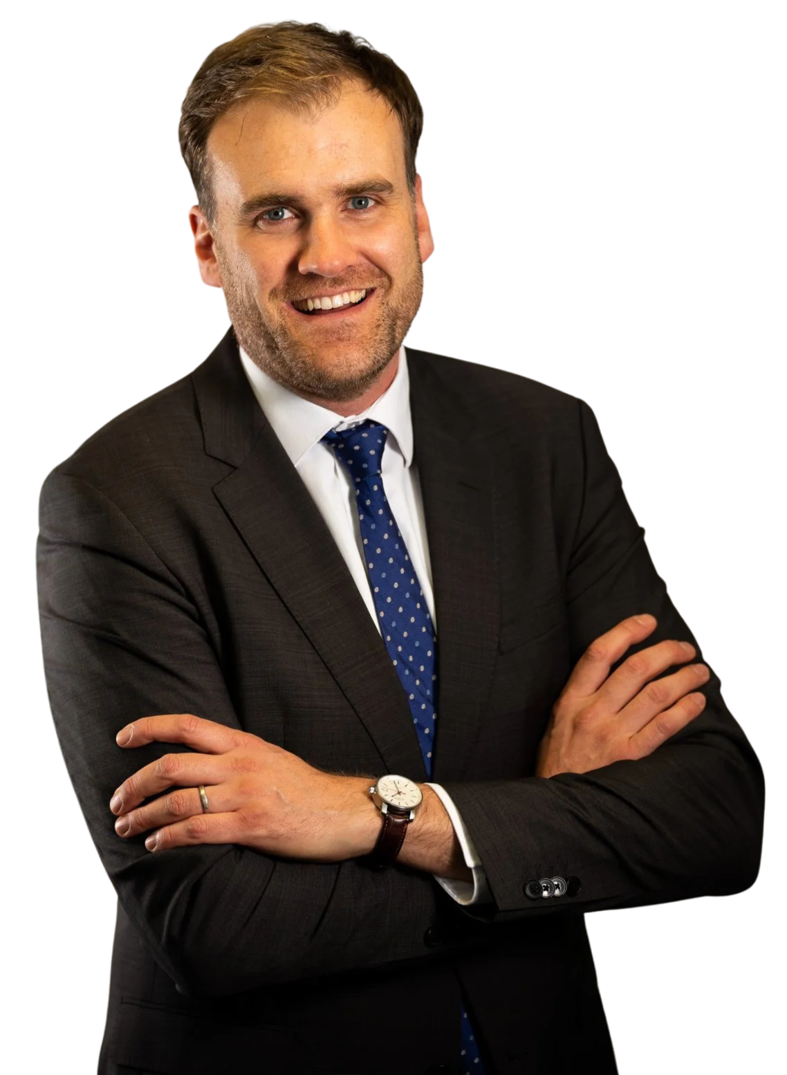 A smiling man with light brown hair, blue eyes, and a beard, wearing a dark suit, white shirt, and blue polka dot tie, with arms crossed, standing against a black background.
