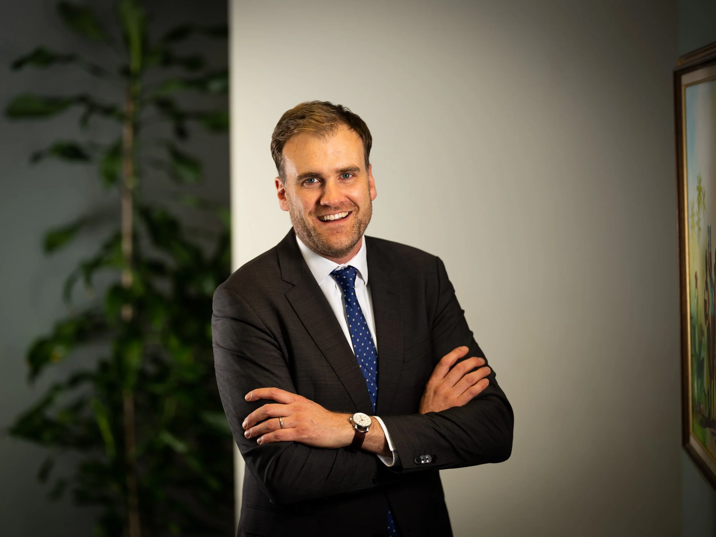 A man in a black suit with a blue polka dot tie, smiling with arms crossed, standing in an office or gallery setting with a plant and framed artwork in the background.