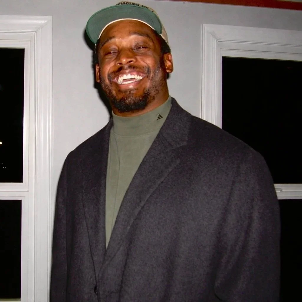 A man with a beard, wearing a green baseball cap, smiling, dressed in a dark blazer and a gray turtleneck shirt, standing indoors near a window.