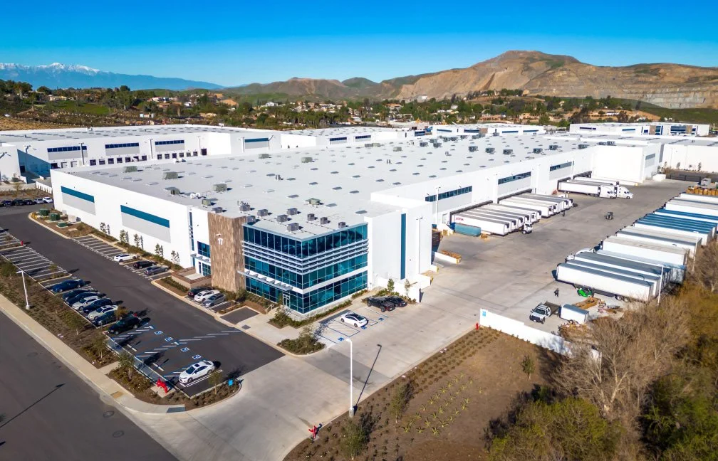 An aerial view of a large modern warehouse and distribution center with parked trucks and cars, surrounded by mountains in the background.