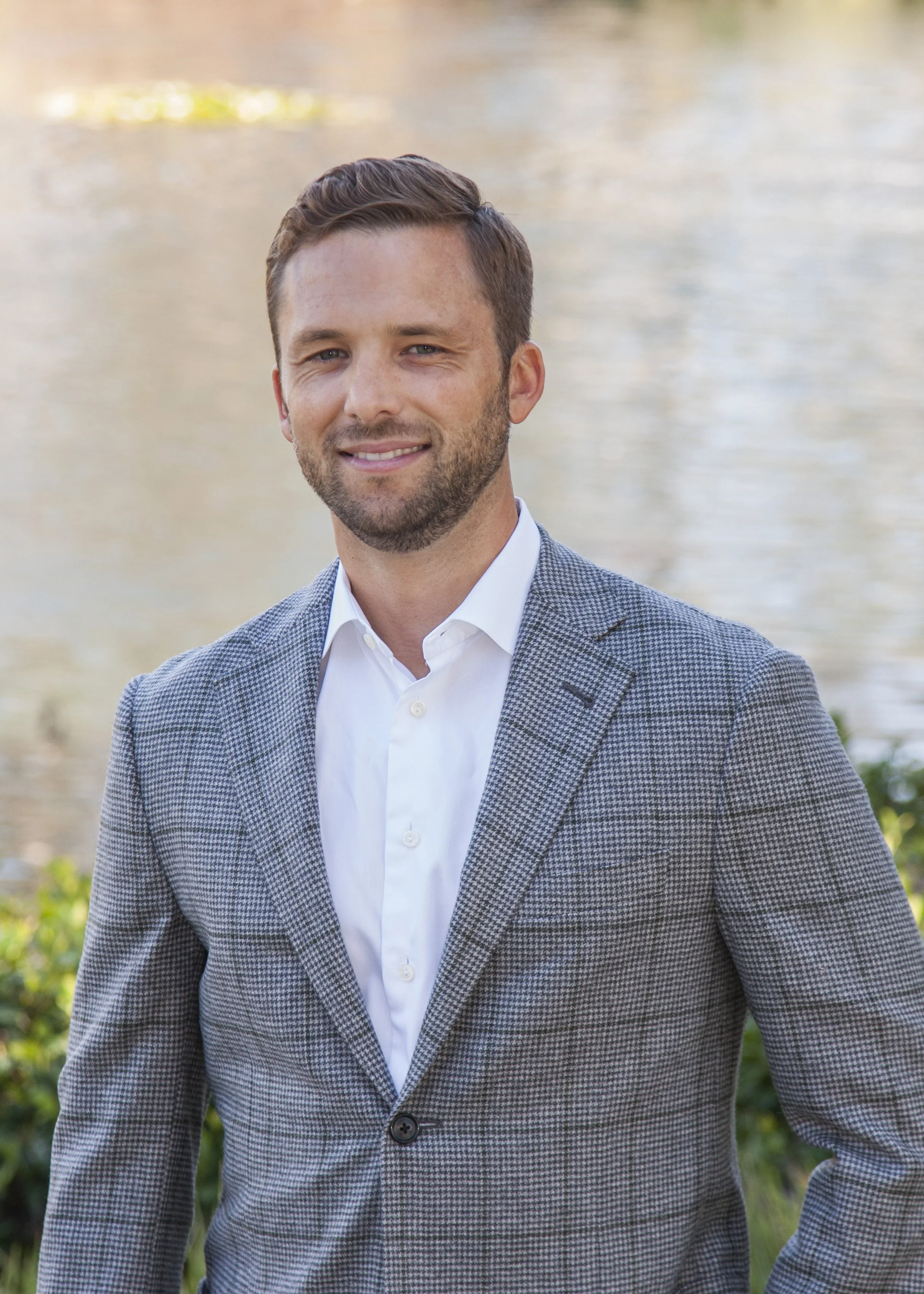 A man with short brown hair and a beard, wearing a light blue checkered blazer and a white shirt, standing outdoors by a body of water with greenery in the background.