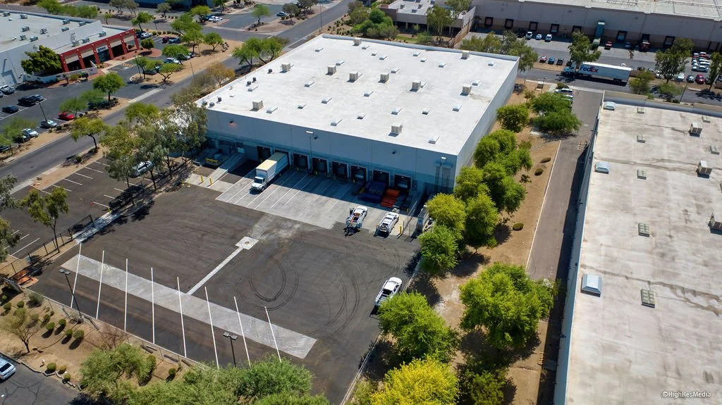 A loading dock area outside a warehouse with several parked vehicles, including a Penske truck and passenger cars. A man is walking towards the building, and a leafless tree is visible in the foreground. Los Angeles cold storage, Orange County