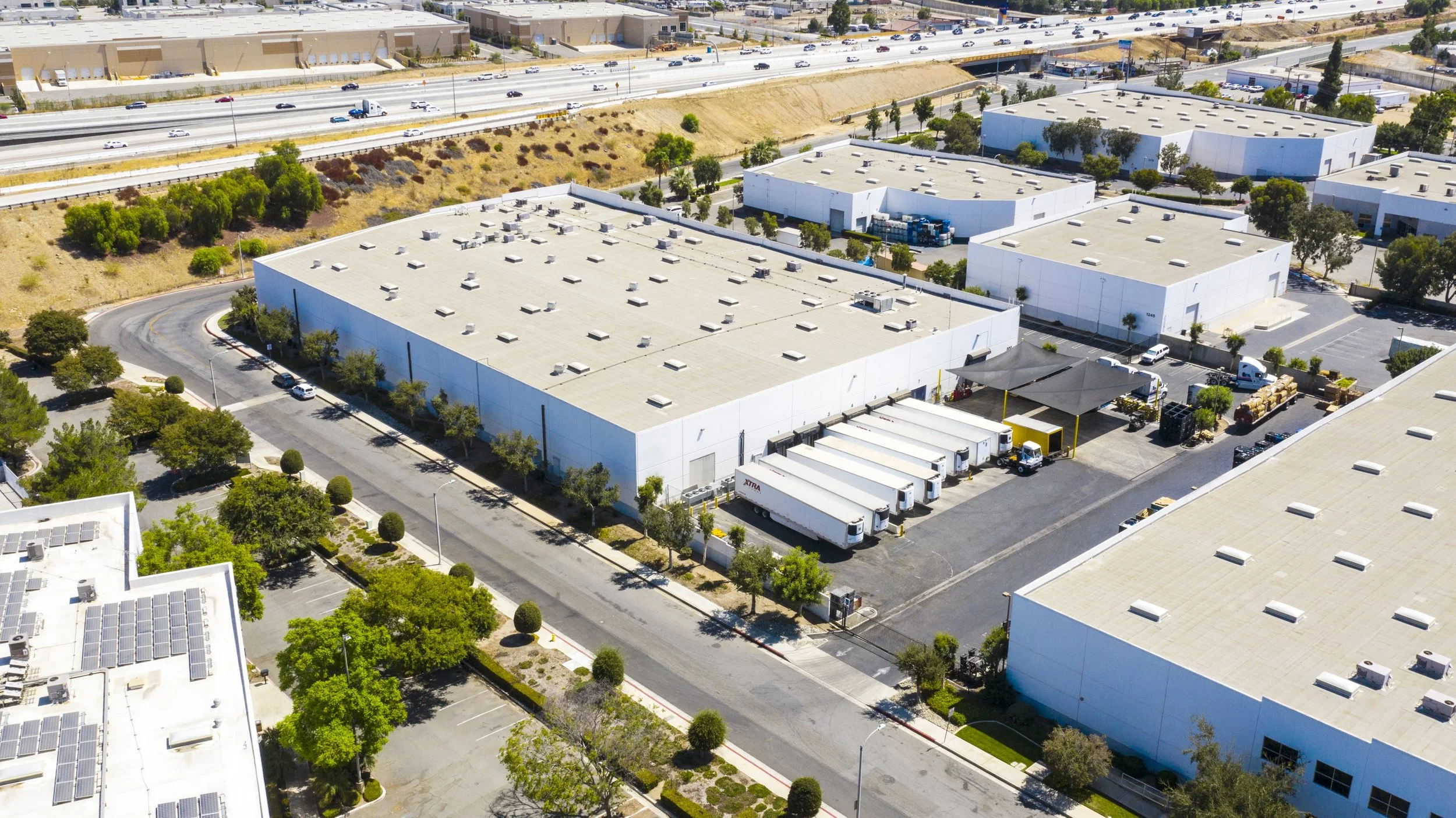 Aerial view of a commercial warehouse complex with multiple large white buildings, parking lots, trees, trucks, and a highway in the background.