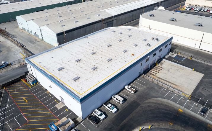Aerial view of a large industrial warehouse with parking lot, some vehicles, and neighboring warehouse buildings. Los Angeles cold storage, Orange County cold storage warehouse, SoCal refrigerated storage, California food warehousing