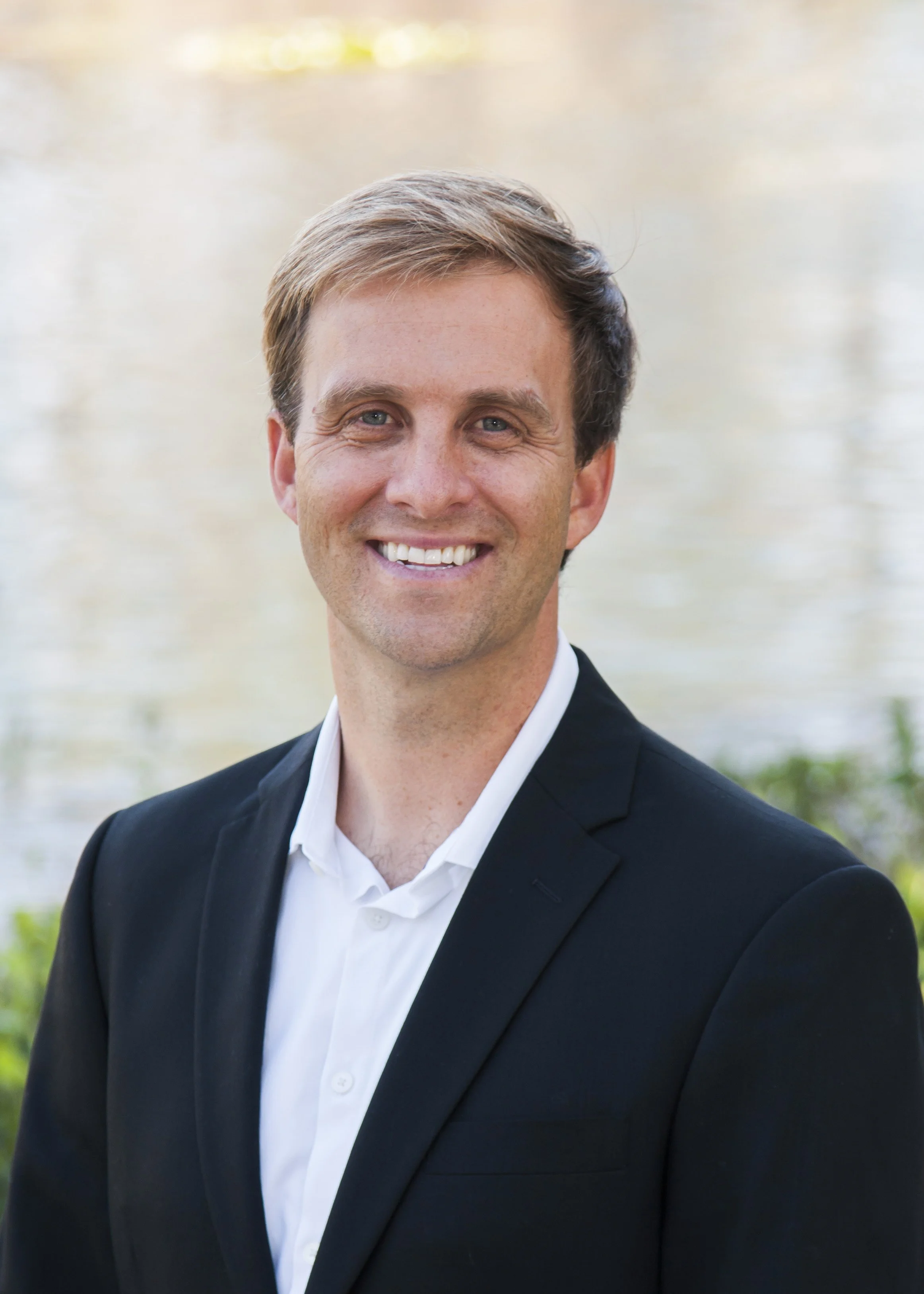 A smiling man in a black suit and white shirt standing outdoors near a body of water with a blurred natural background.