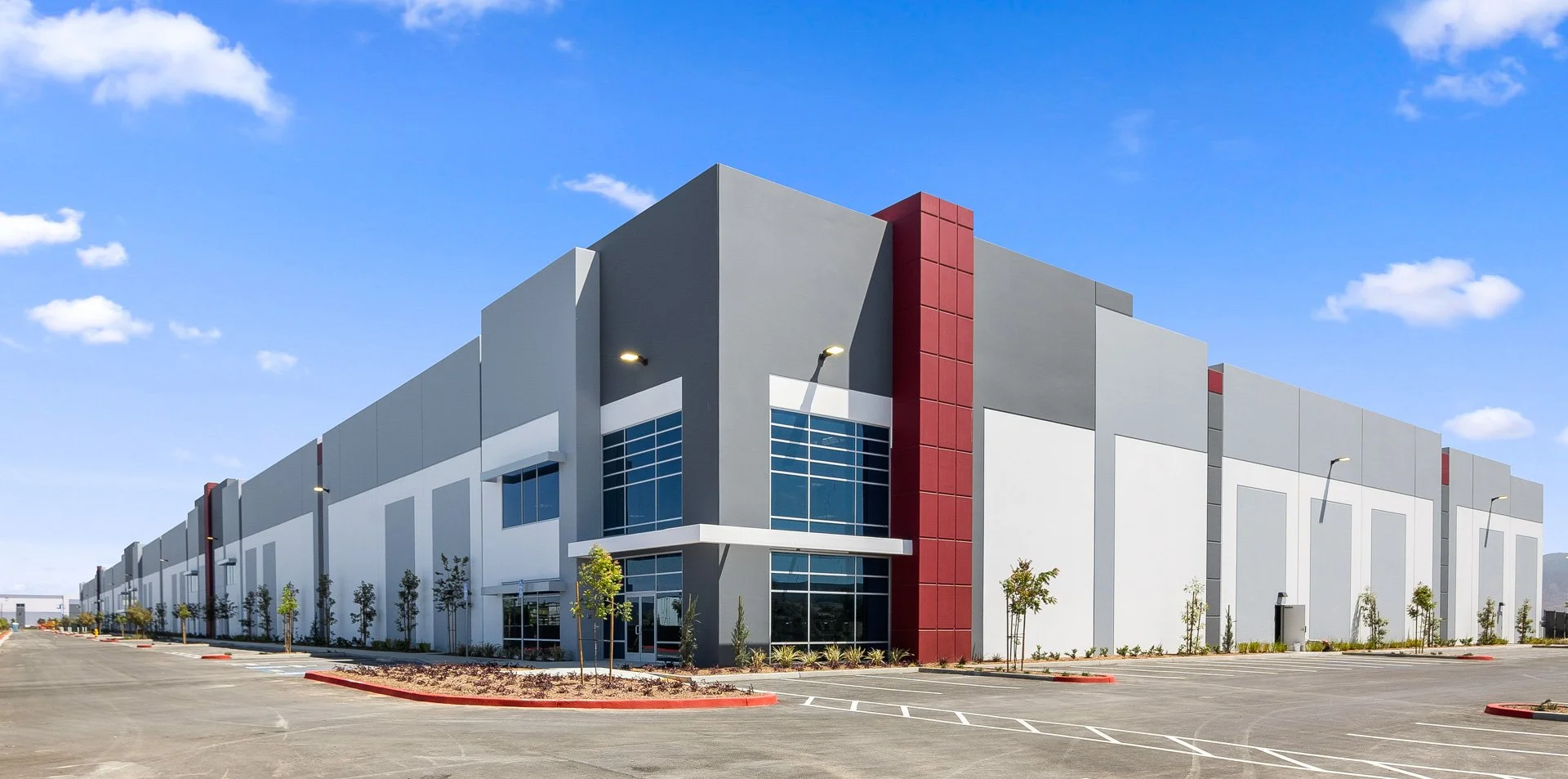 Large modern warehouse with a mostly gray facade, red accents, and large windows, surrounded by a parking lot with small trees and clear blue sky.