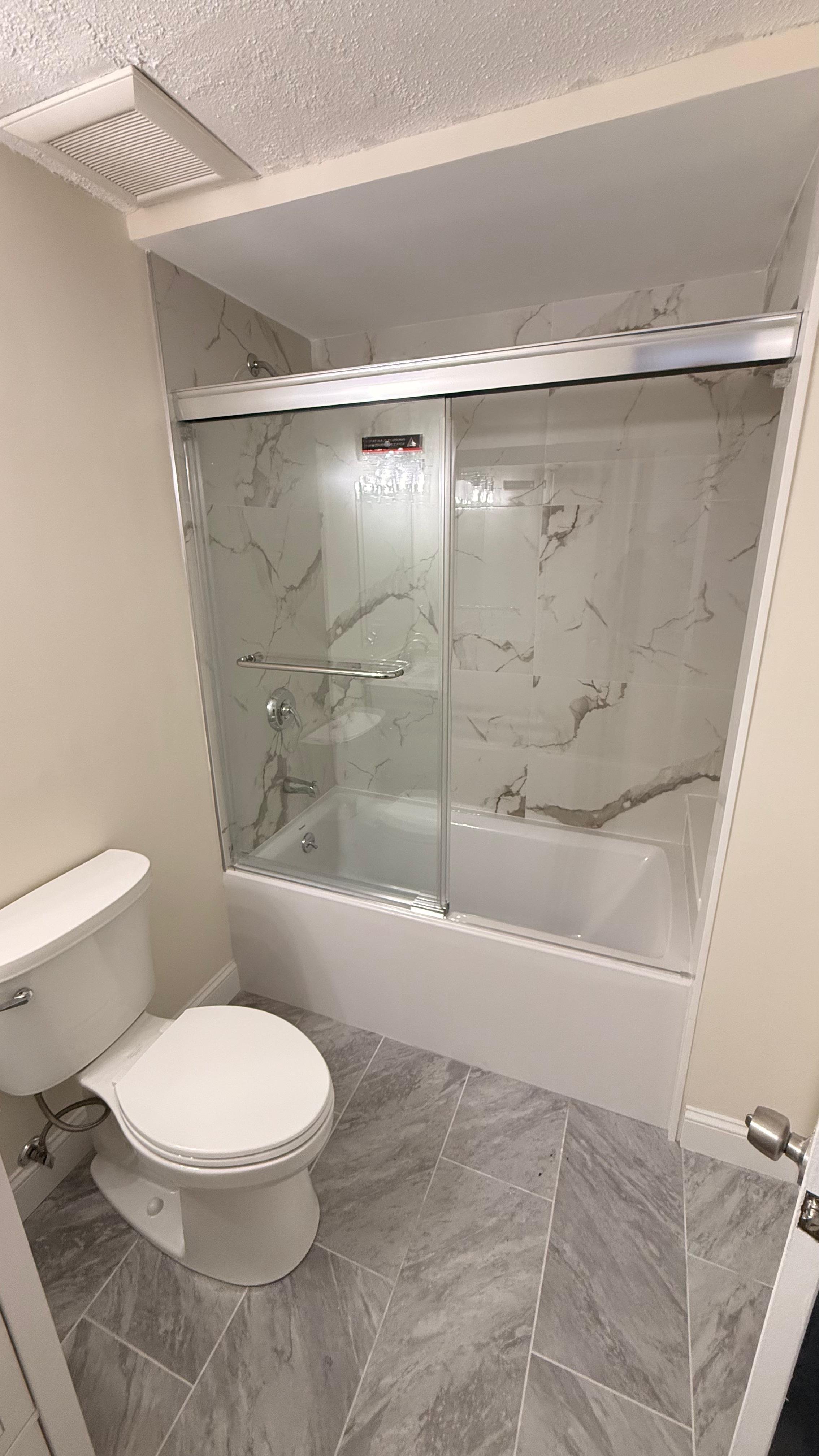 Bathroom with a toilet in the foreground and a bathtub behind a glass sliding door with marble pattern walls and grey tiled floor.