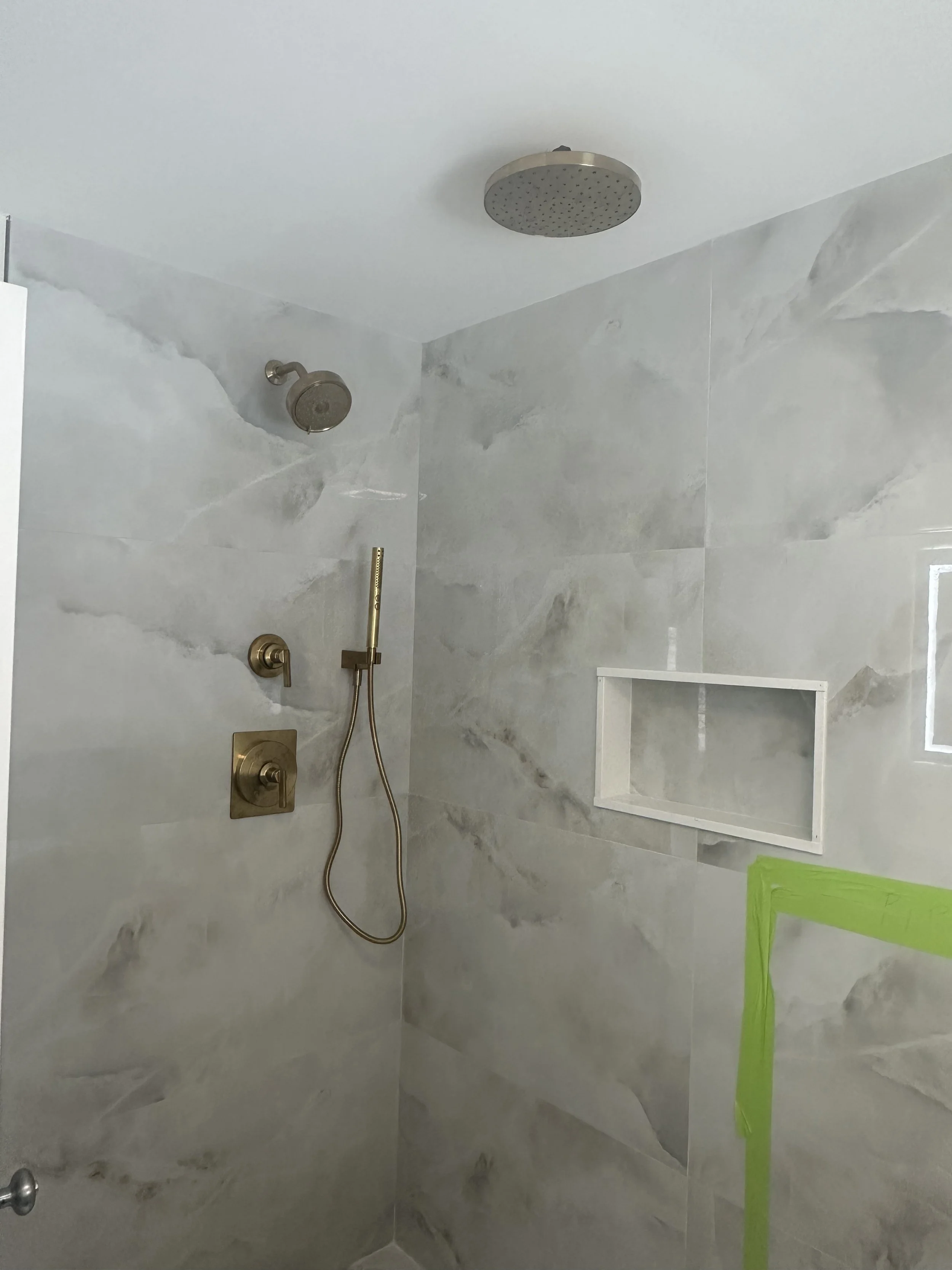 Bathroom shower with large round showerhead at ceiling, bronze handheld shower attached to wall with handle, built-in shelf in the tiled wall, partially taped corner near the lower right.