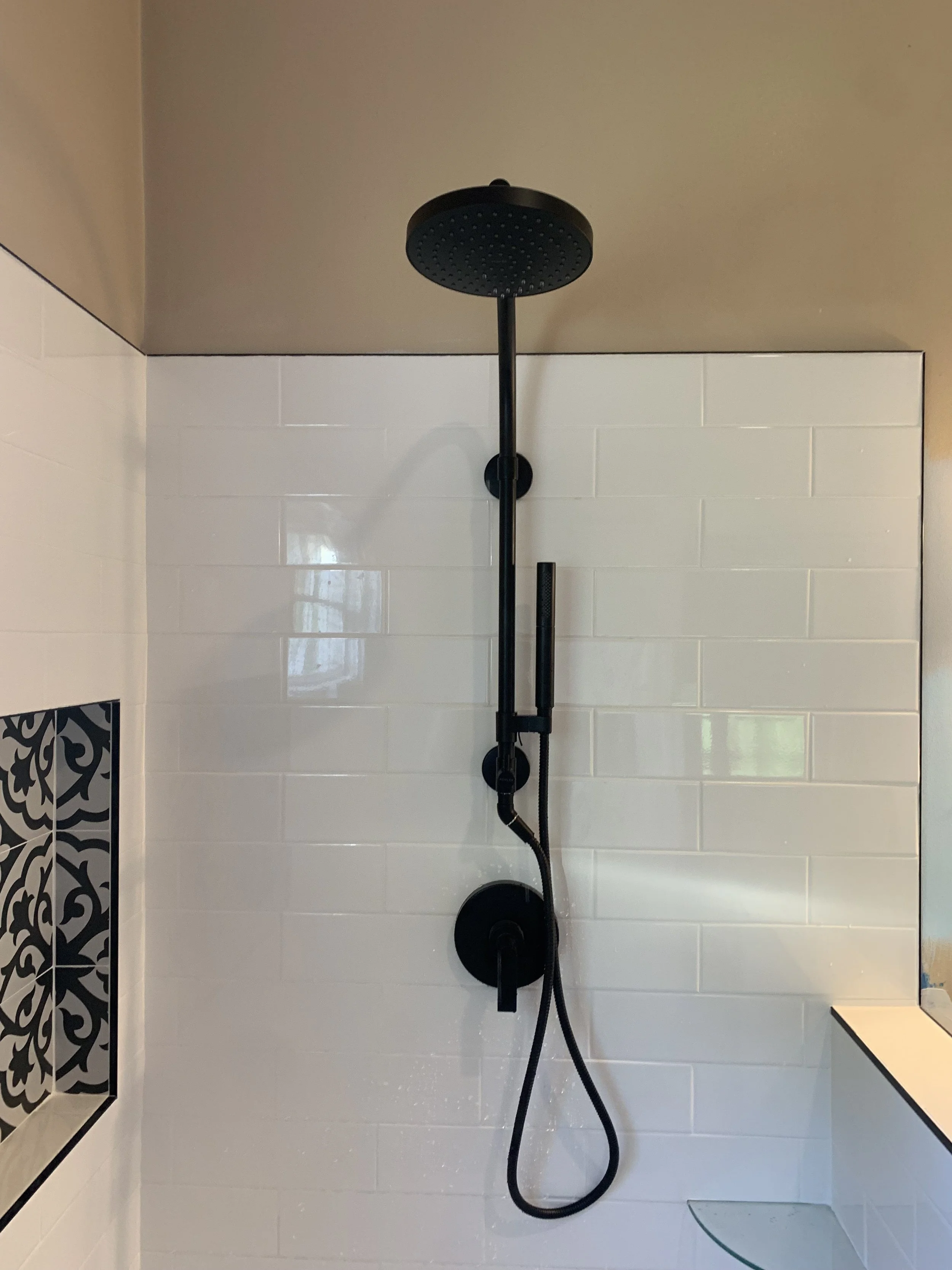 Black showerhead, handheld shower, and hose mounted on a white tiled bathroom wall with some decorative tiles and a small section of a glass shelf.