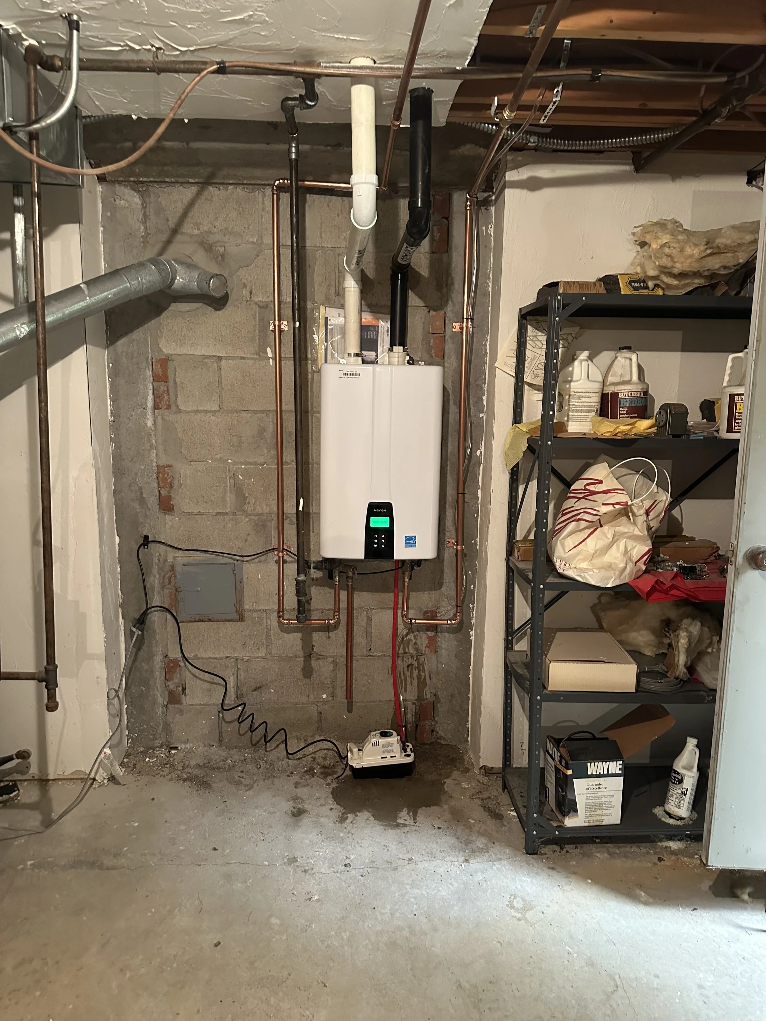 A utility room with an industrial boiler, copper pipes, a shelf with cleaning supplies, and various tools and boxes on the floor.