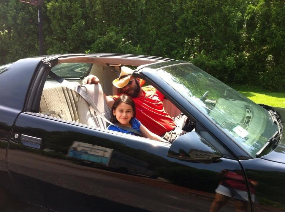 A man and a young girl sitting inside a black convertible car parked outdoors, with green trees in the background. The man is wearing a cowboy hat and sunglasses, smiling, while the girl is smiling at the camera.