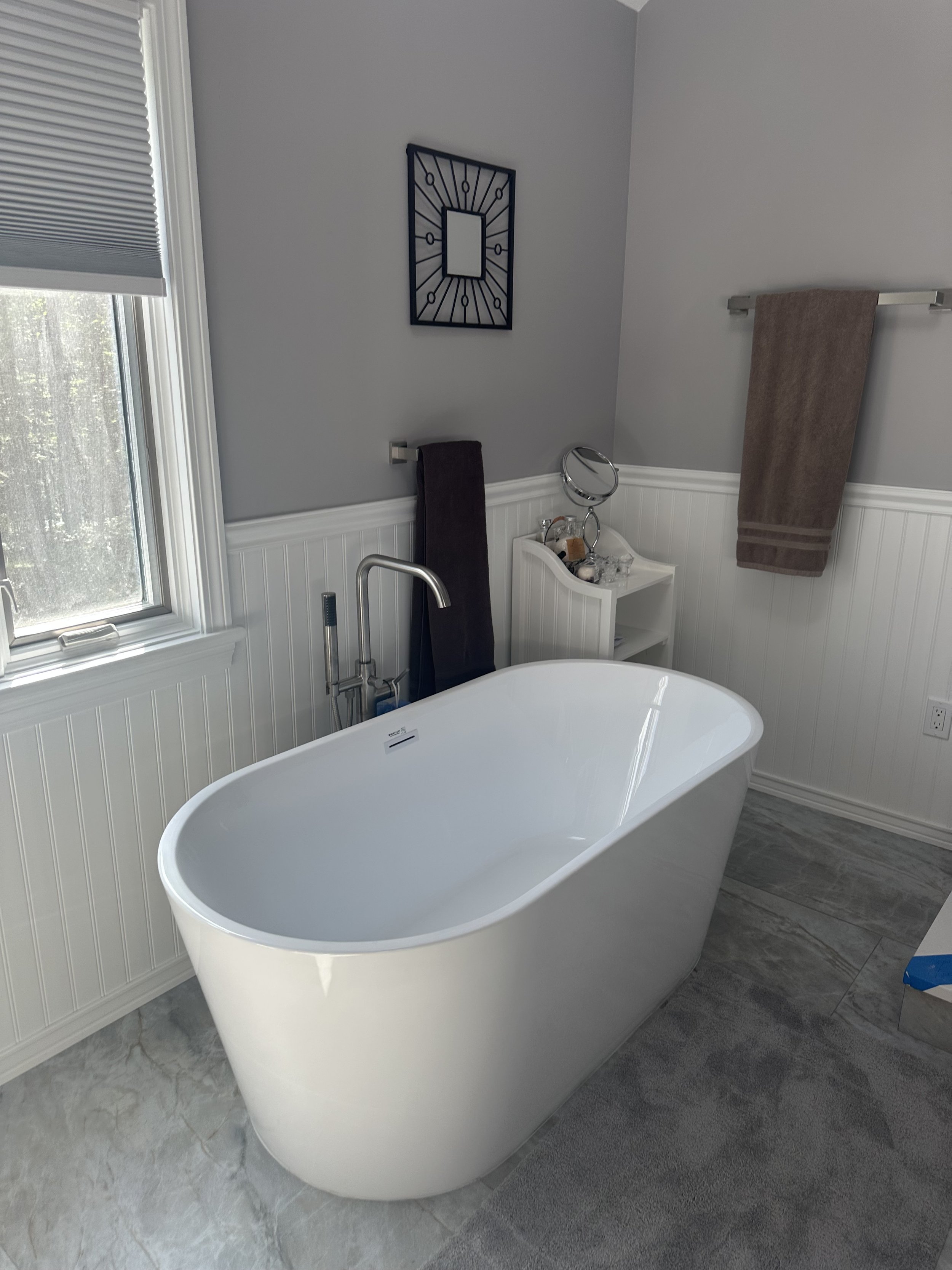 Modern bathroom with freestanding white bathtub, gray floor tiles, gray painted walls, a window with blinds, towel racks with brown towels, and wall art.
