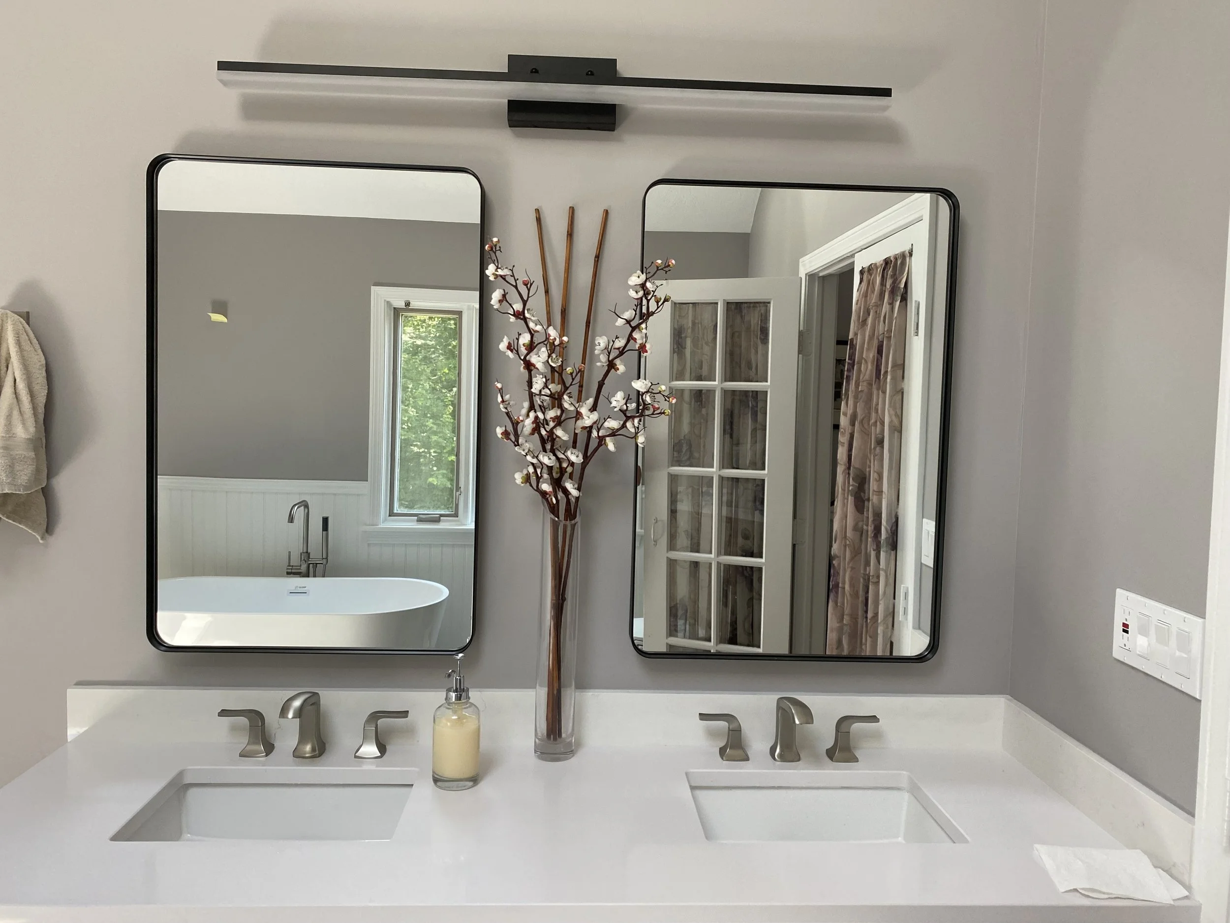 A bathroom mirror with a white countertop, a modern faucet, a soap dispenser, and a clear glass with flowers. Two black-framed mirrors hang above the sink, reflecting a window and part of the bathroom interior.