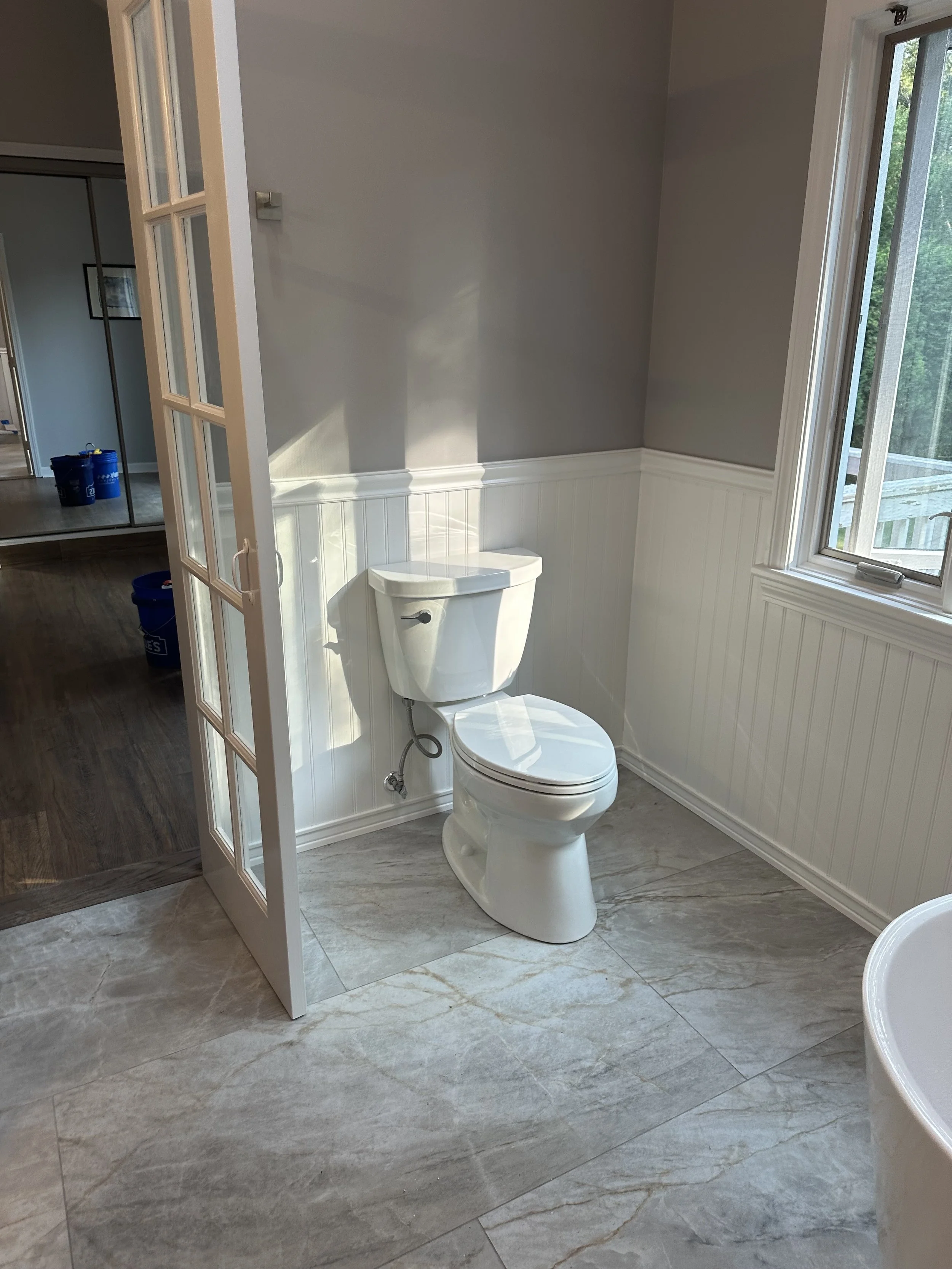 A bathroom with gray and white marble floors, a window with a view of trees, and a toilet. A white wainscoting runs along the wall, and there's a glass door with a wooden frame separating the bathroom area from another room.