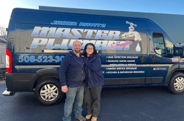 A man and woman standing in front of a large blue service van labeled "Master Plumbing & Drain" with a faucet graphic, phone number, and services listed.
