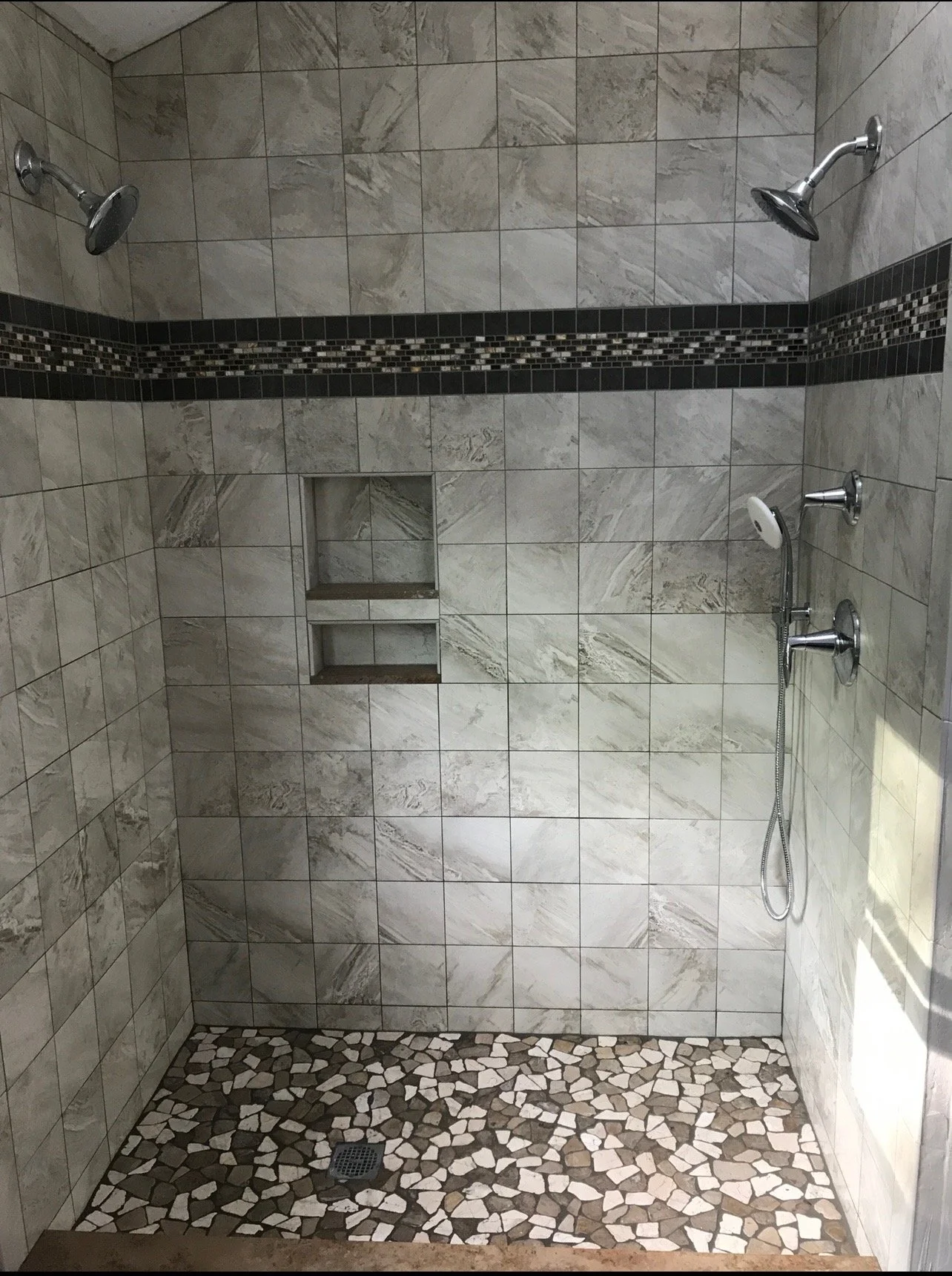 A tiled shower with two shower heads, a small built-in shelf, and a pebble floor.