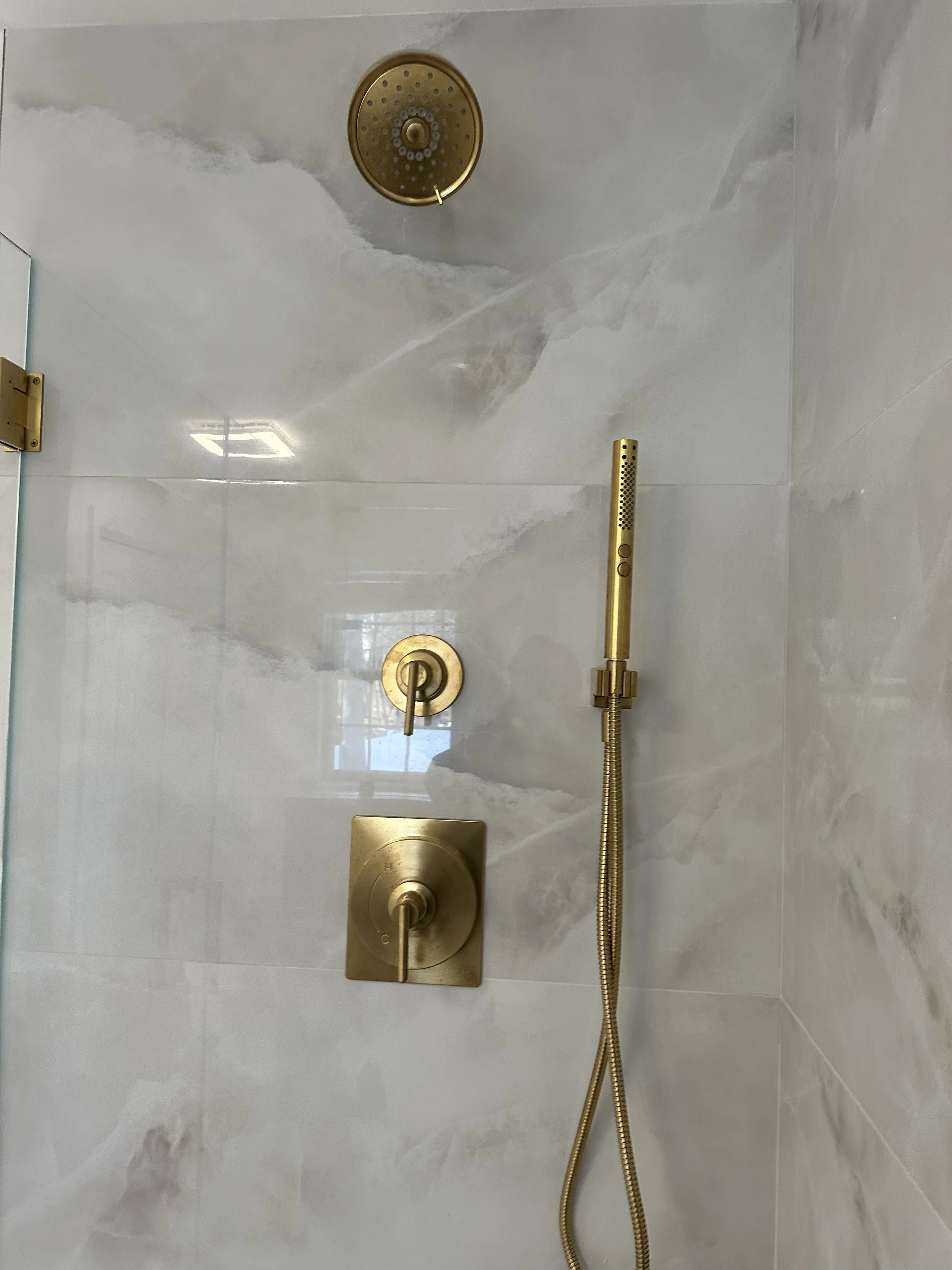A gold-colored showerhead, a handheld shower wand, a round temperature control handle, and a square diverter knob on a marble wall shower.